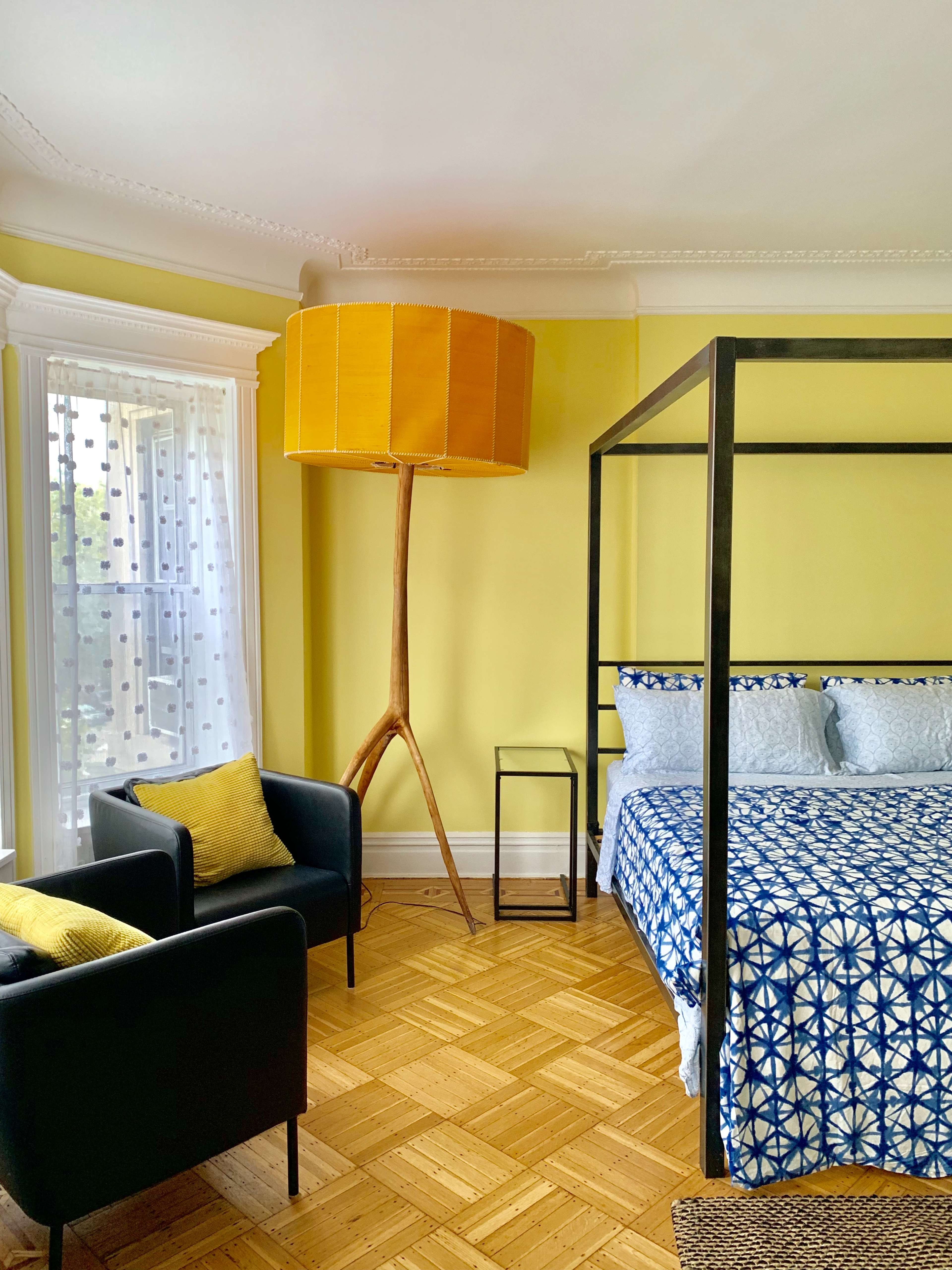 The room features a yellow-walled space with a four-poster bed, a tall yellow lamp, and two black armchairs.