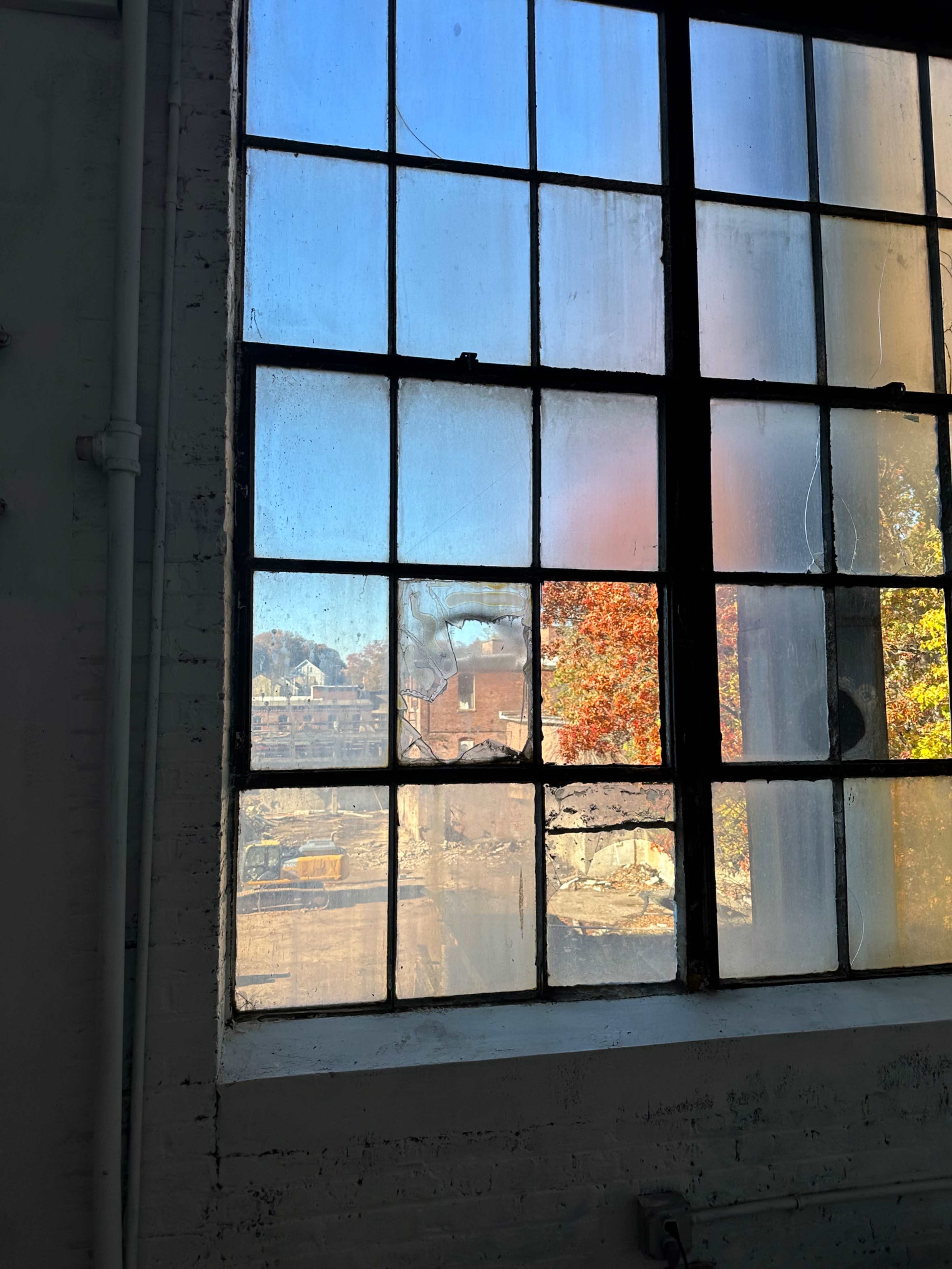The image shows a large, industrial window with several cracked panes, through which a vivid autumn scene is visible outside.