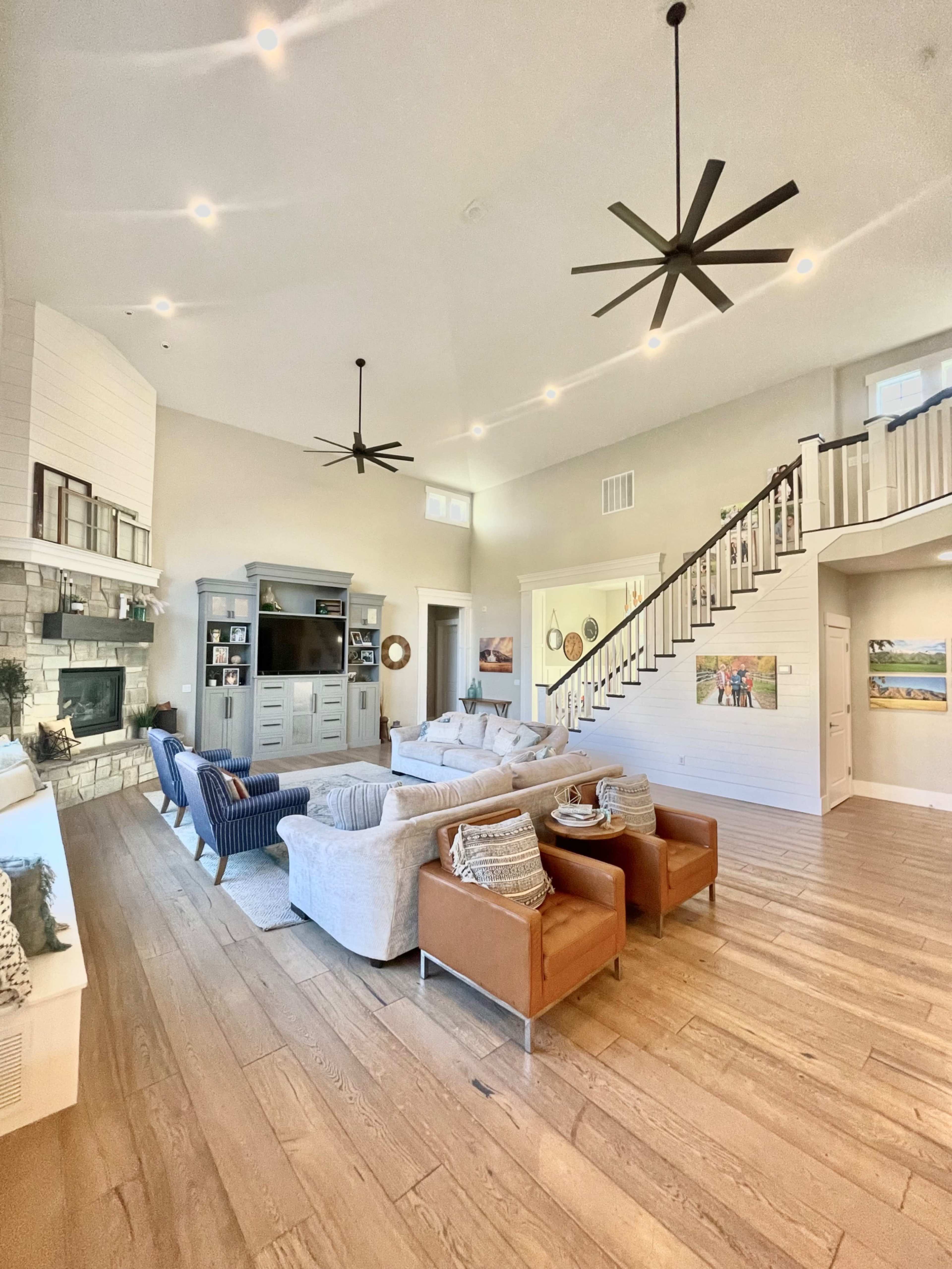 A spacious living room with a large sectional sofa, two armchairs, and a television set against a light-colored wall, illuminated by several ceiling fans and recessed lighting.