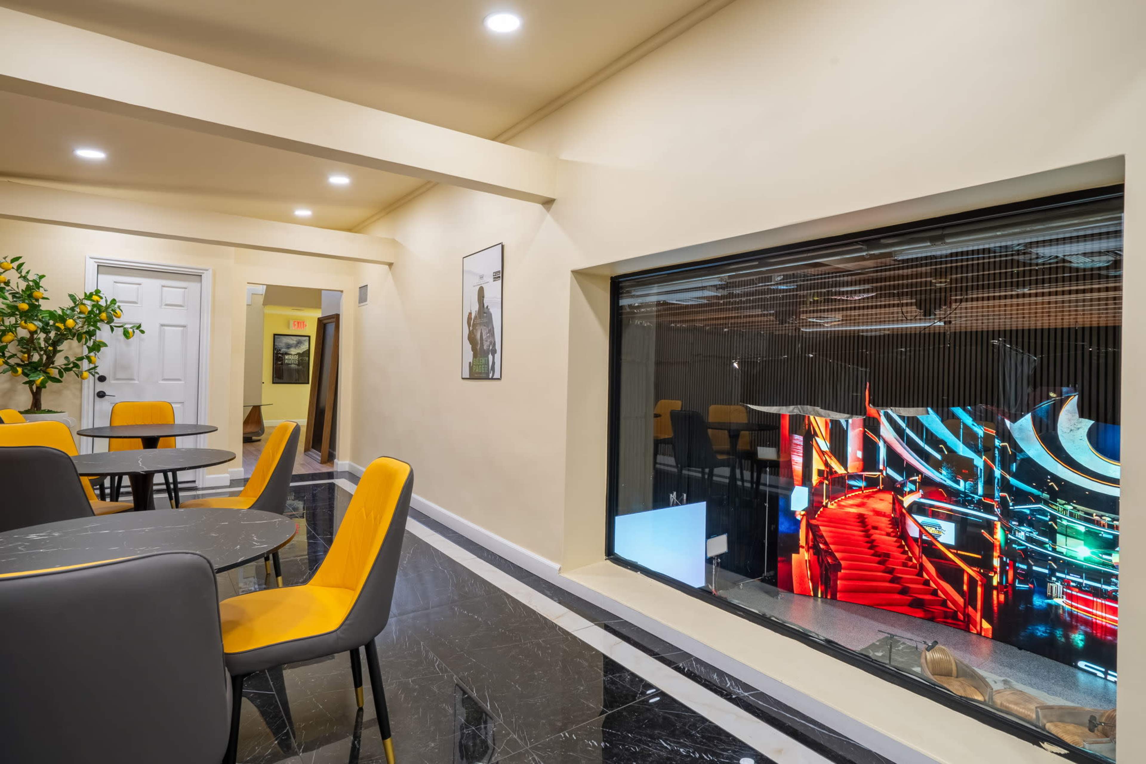 The image shows a modern interior space with yellow and gray dining chairs around black marble tables, and a large window displaying a colorful, illuminated staircase scene.