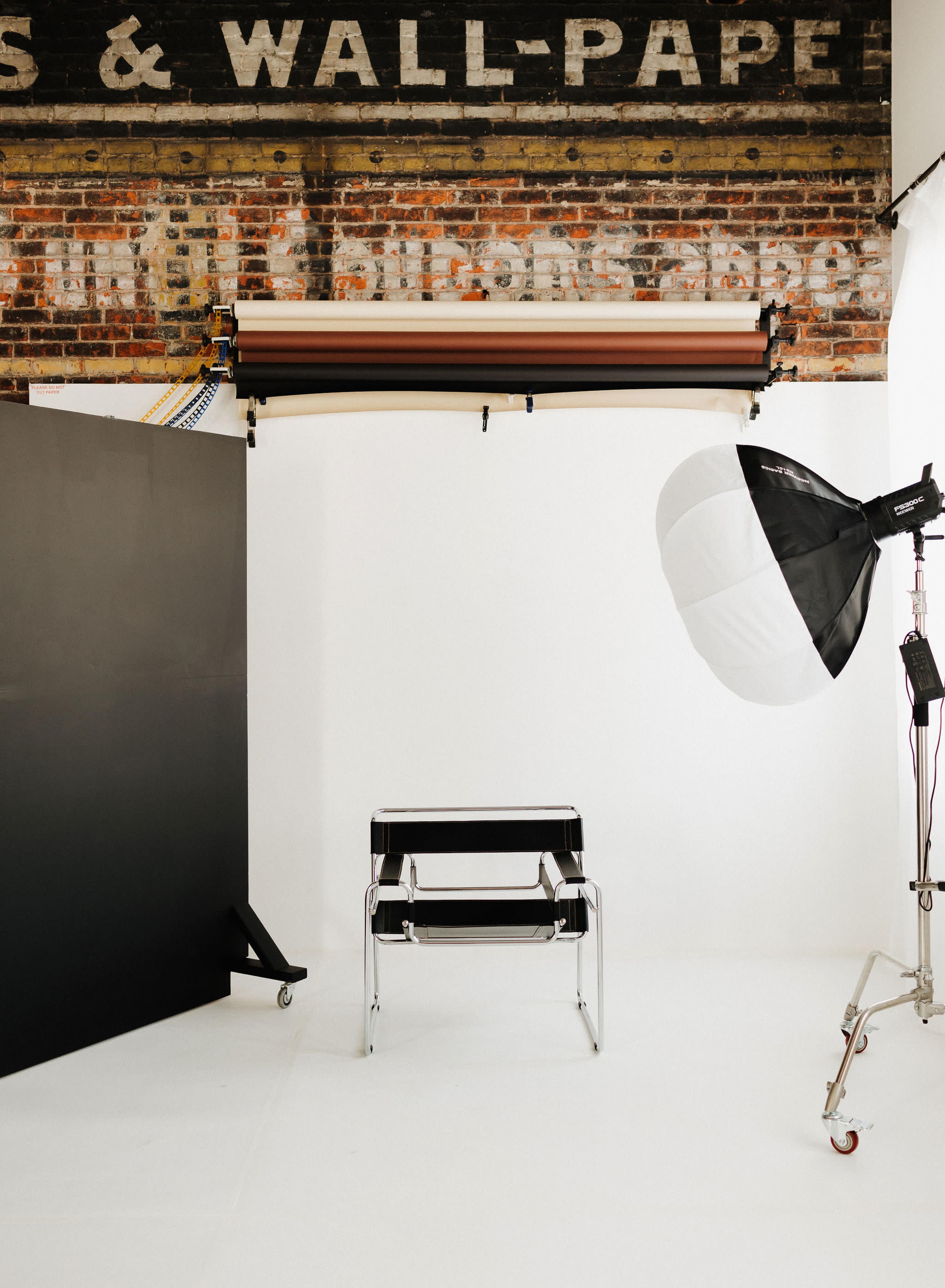 The image shows a minimalist photography studio setup with a black chair, a light source, and a backdrop against a brick wall.