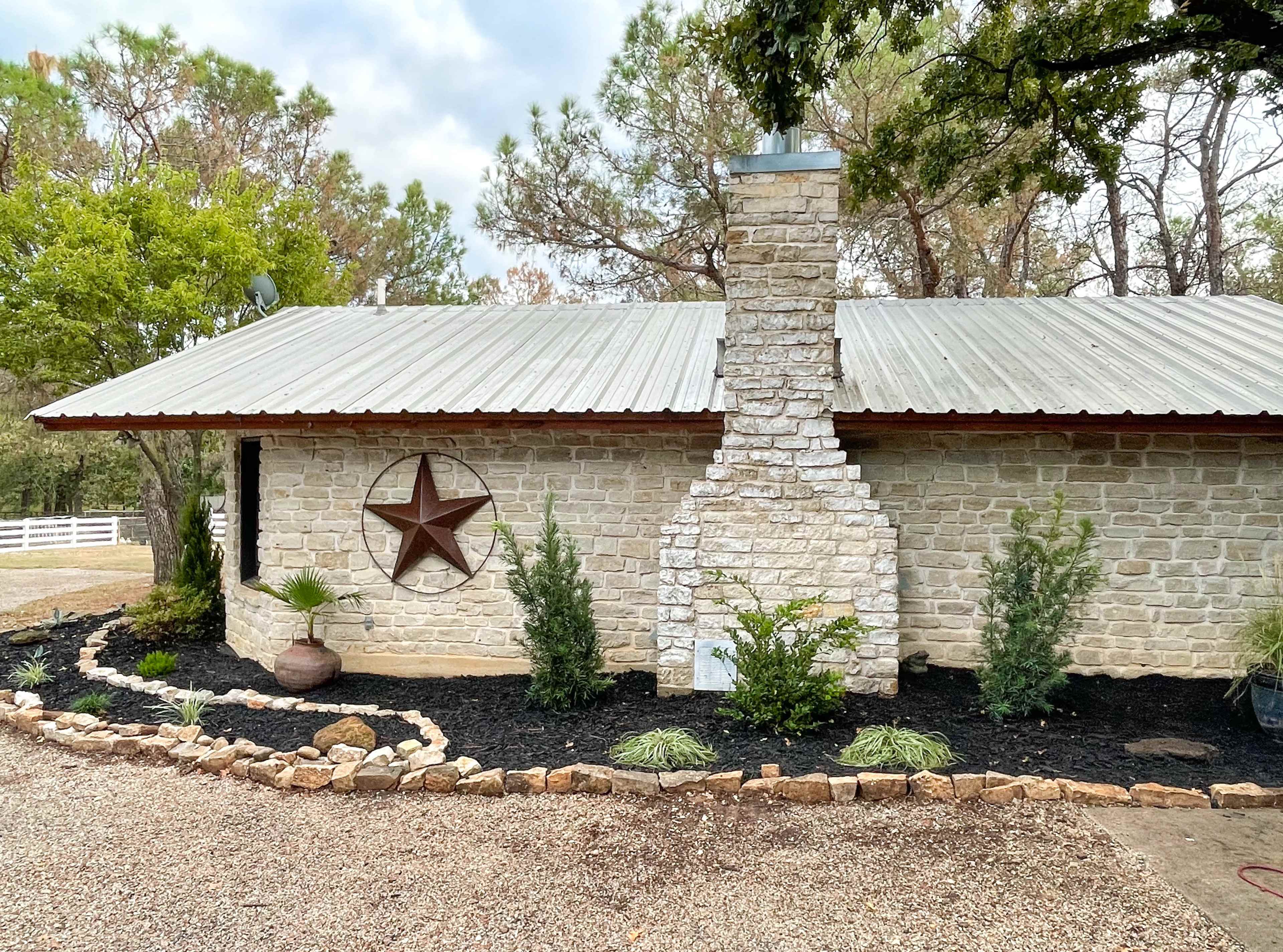 A stone cottage with a metal roof, featuring a central chimney and a decorative star, surrounded by landscaped shrubs and gravel.