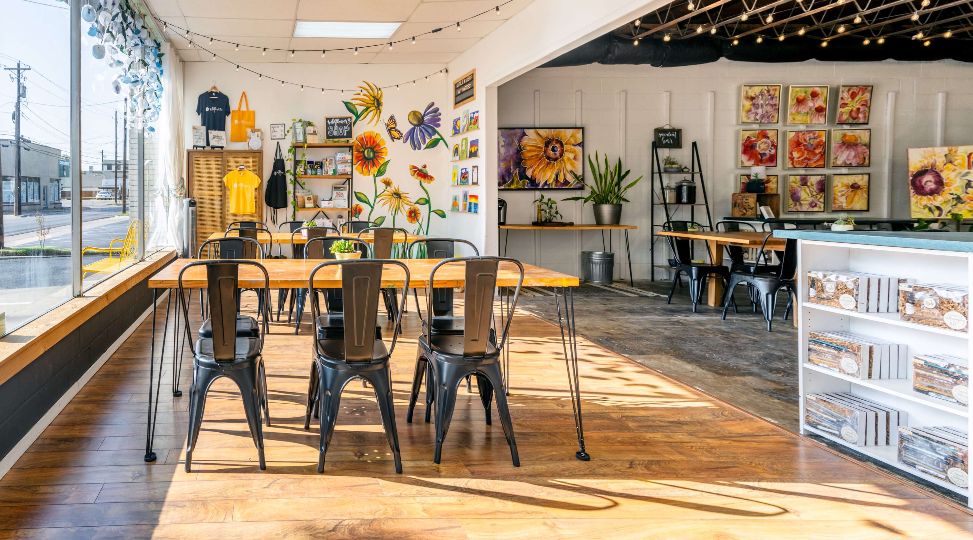 The image shows a brightly lit art studio or café featuring wooden tables, black chairs, and colorful flower artwork on the walls.