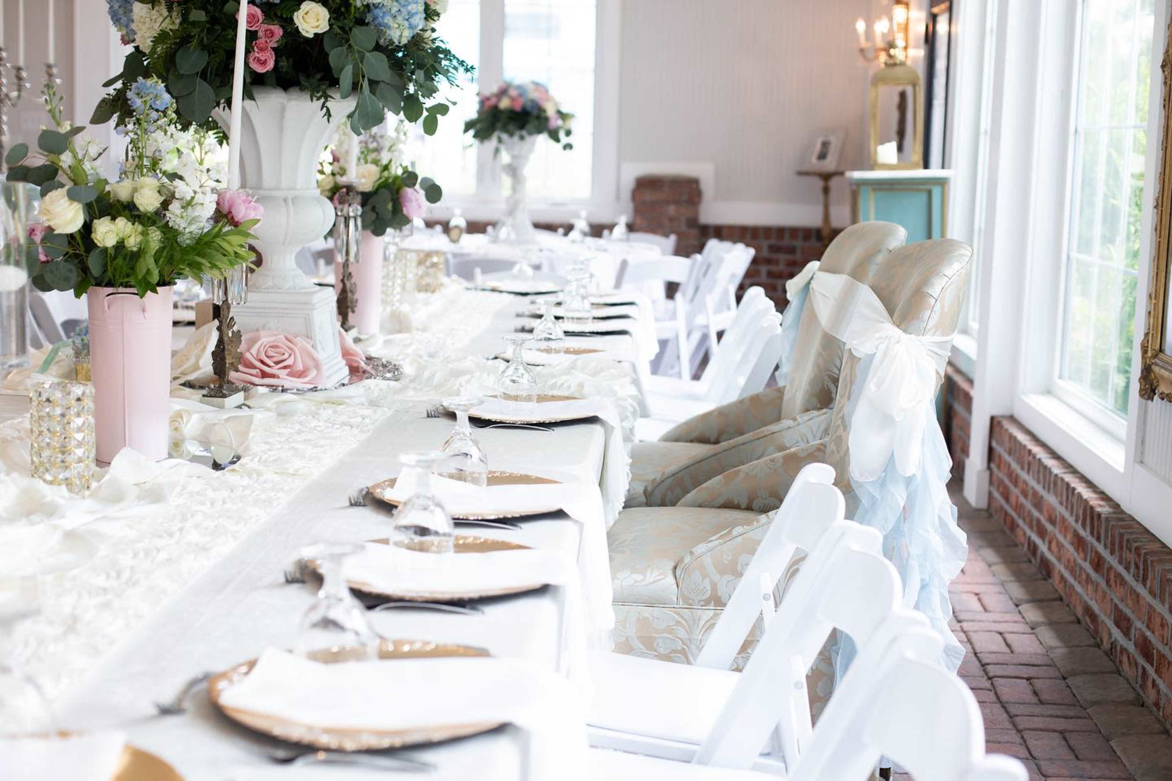 A long dining table is elegantly set with white chairs and a floral centerpiece, surrounded by softly colored decorations.