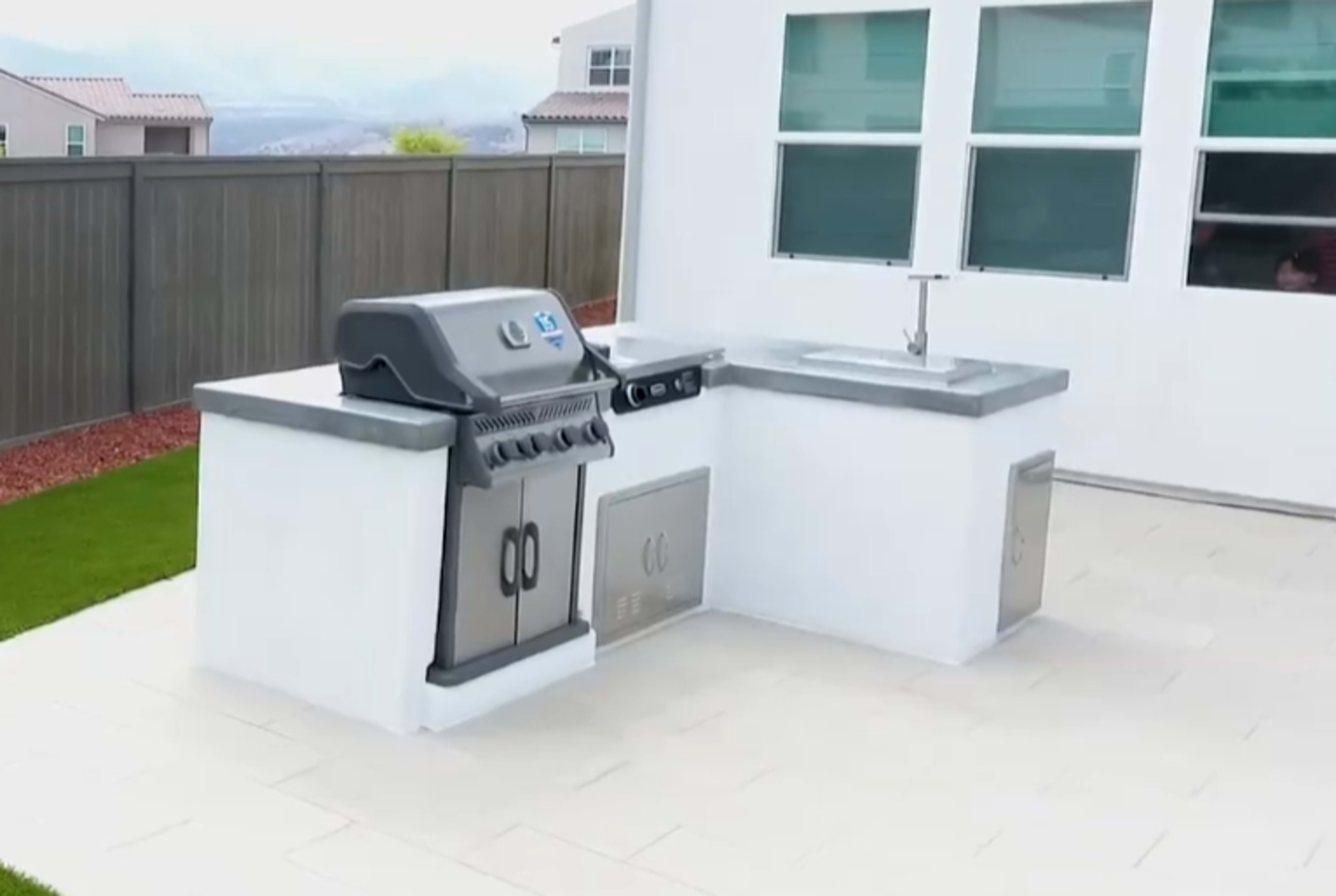 The image shows an outdoor kitchen area featuring a gas grill, a stainless steel sink, and a countertop, set against a backdrop of a fenced yard.