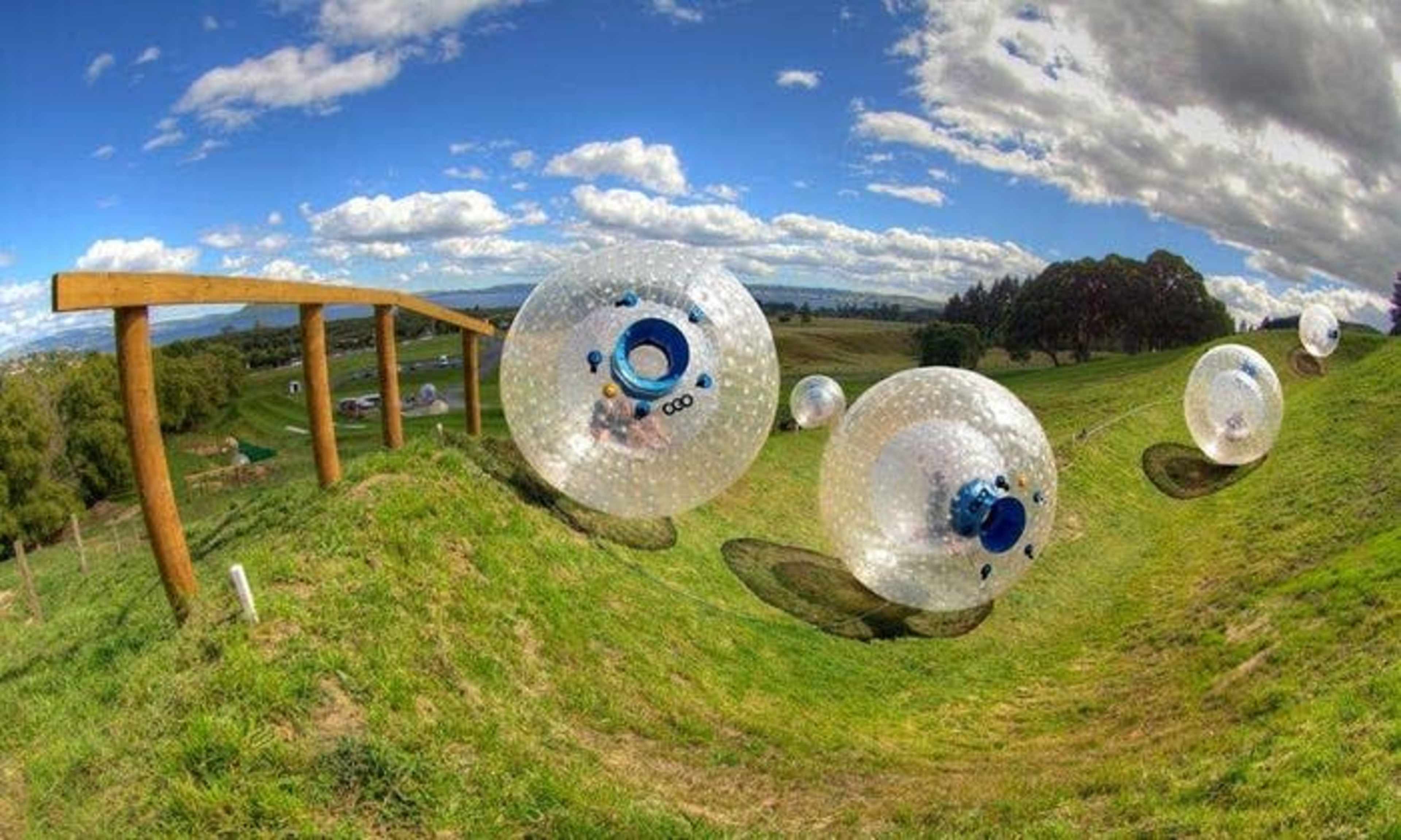 Transparent spheres roll down a grassy hill along a wooden track under a blue sky with clouds.