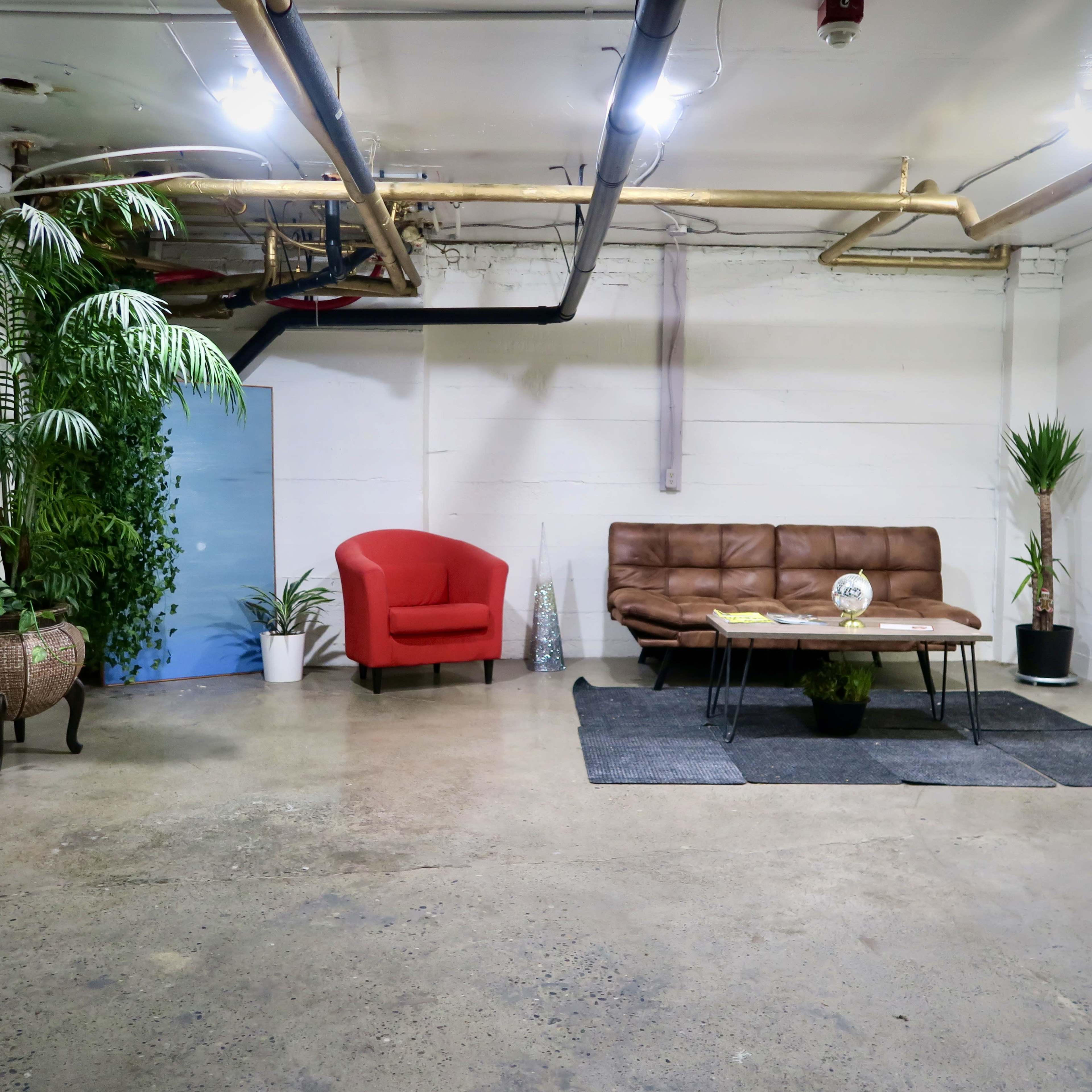 The image shows a minimalist interior space with a red armchair, a brown sofa, a coffee table, and several indoor plants against a plain white wall.