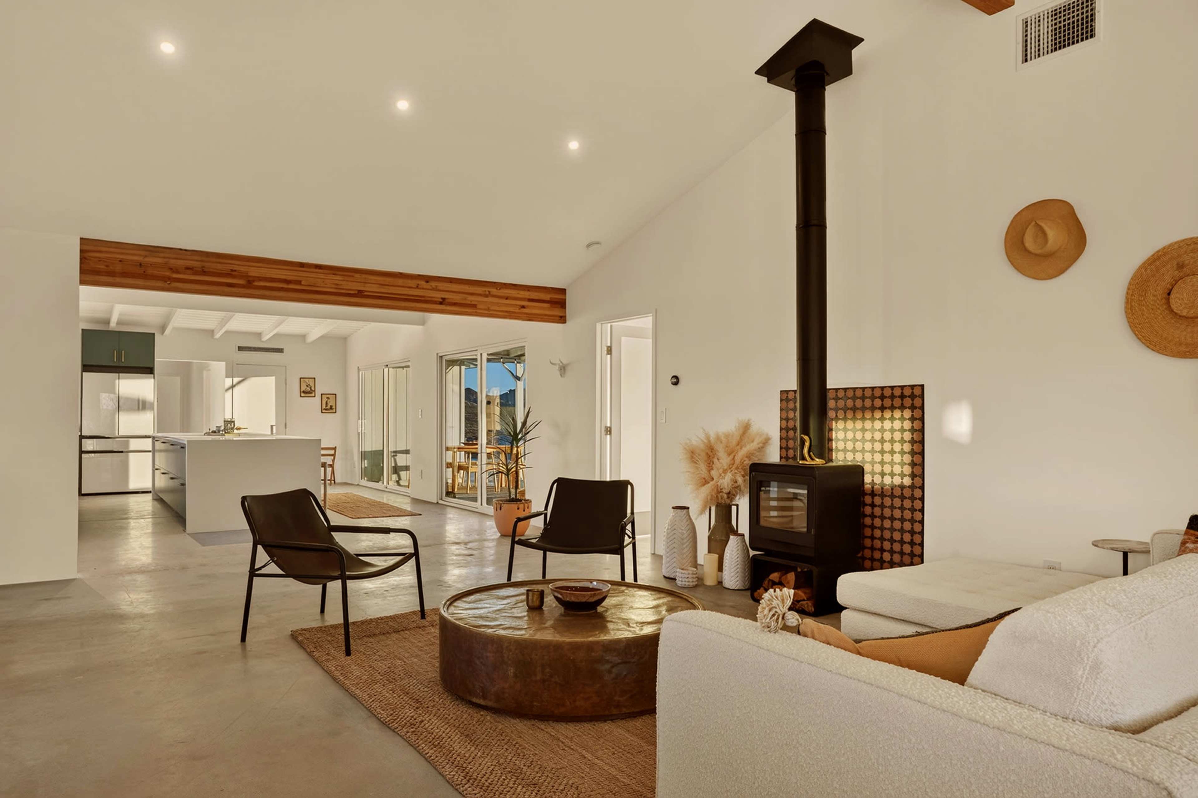 The image shows a spacious, modern living area featuring a circular coffee table, a cozy couch, and a wood-burning stove, with large windows providing natural light.