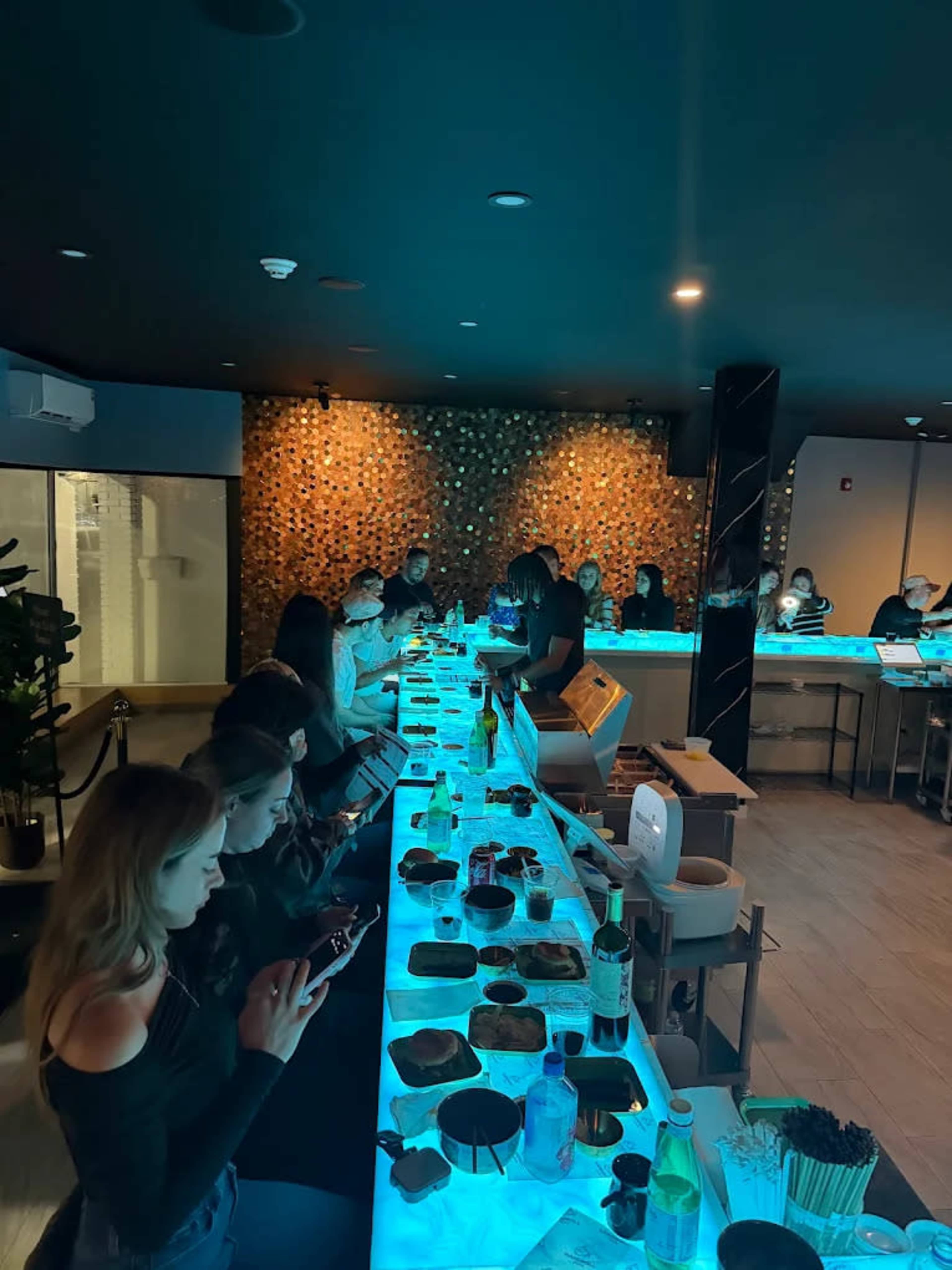 A bar area with patrons sitting at a glowing blue countertop, engaging with their phones while various items are placed on the surface in front of them.