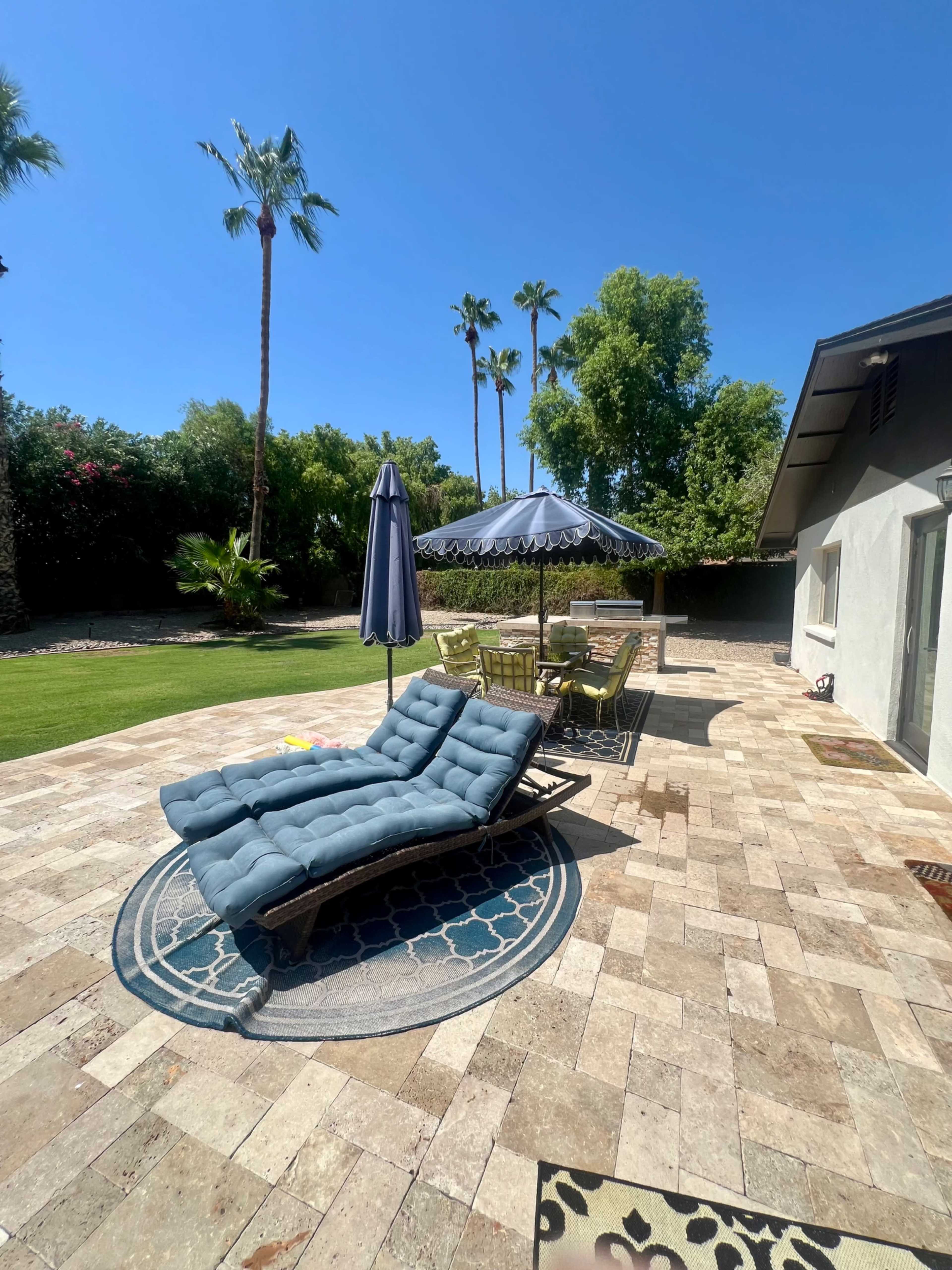 A patio area features a blue lounge chair, a shaded dining table, and palm trees in the background under a clear blue sky.