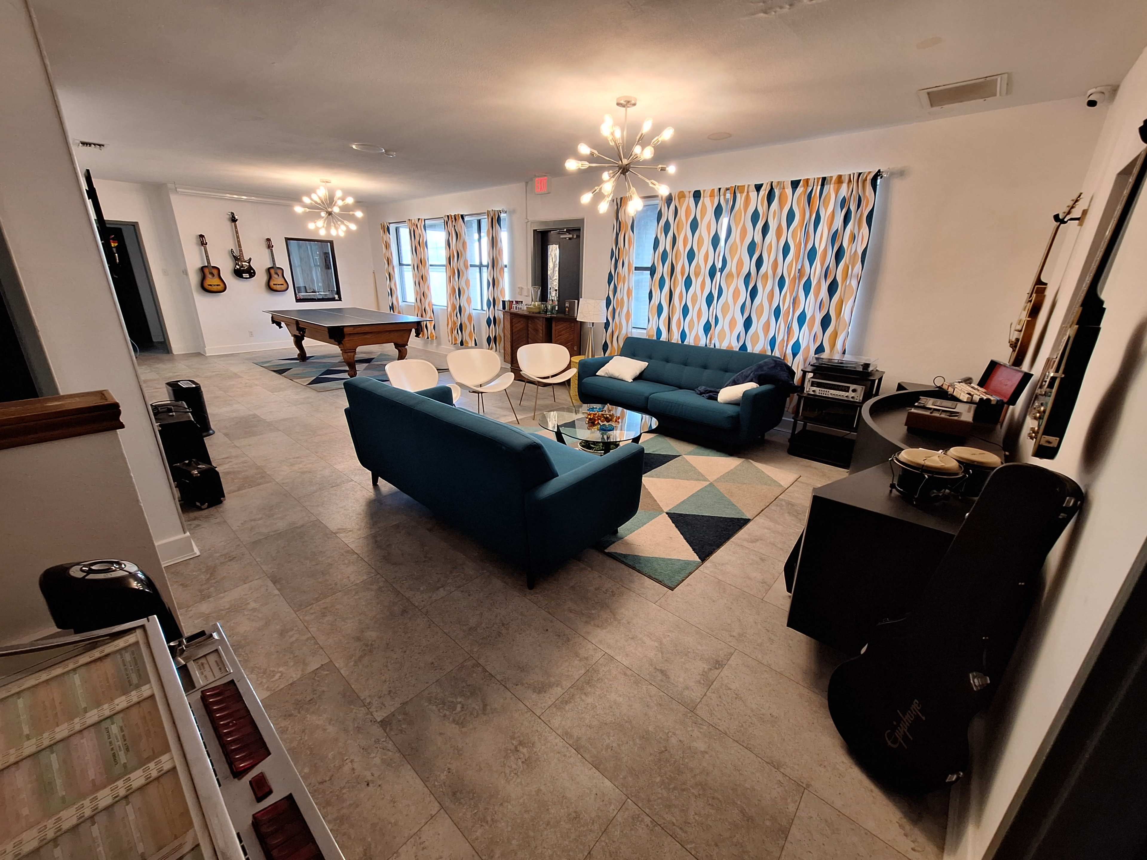 A modern living room features blue couches, a pool table, and musical instruments against a backdrop of patterned curtains and tile flooring.