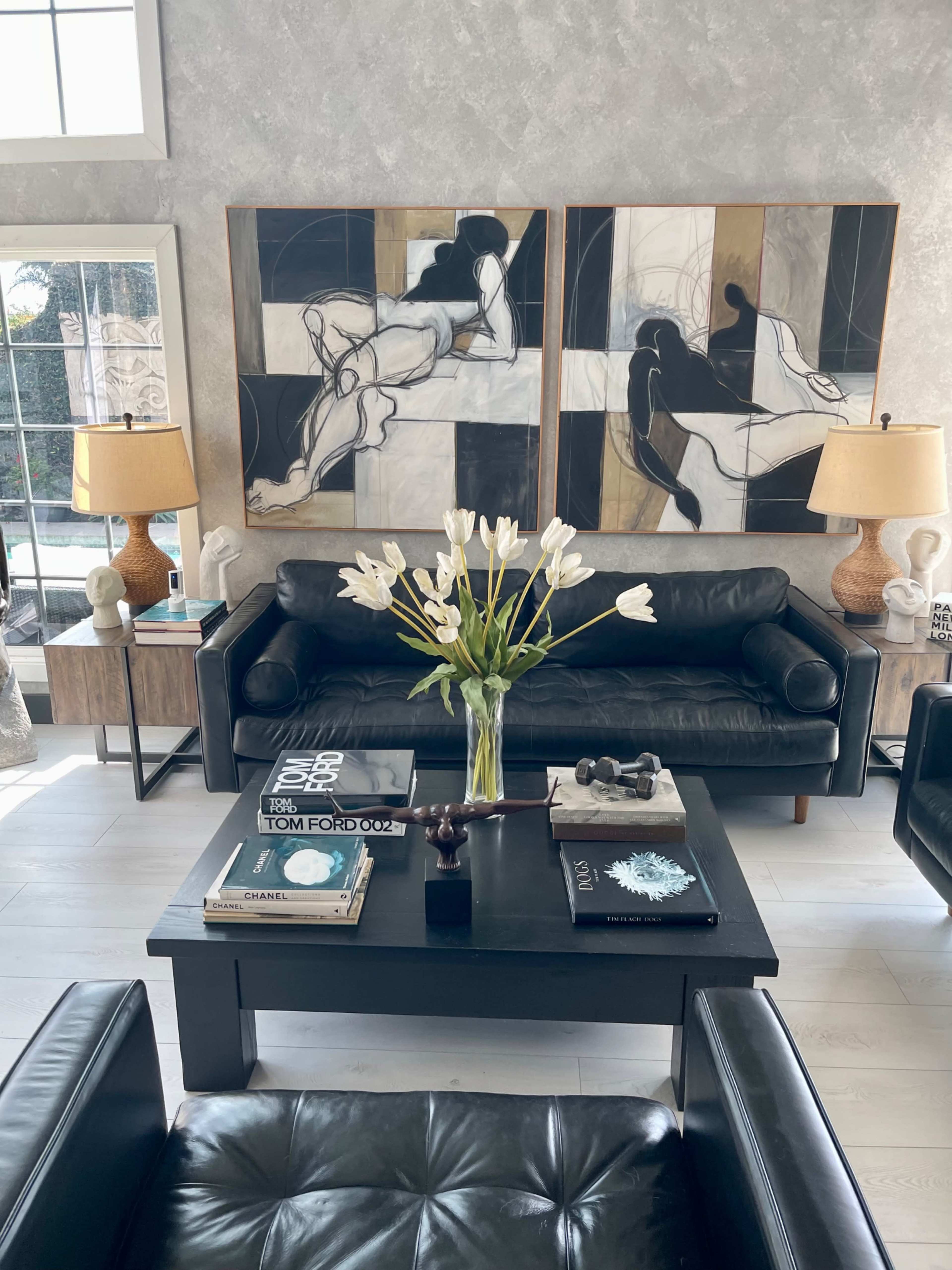 The image shows a modern living room with a black leather sofa, a coffee table adorned with books and a decorative piece, two abstract paintings on the wall, and side tables with lamps.
