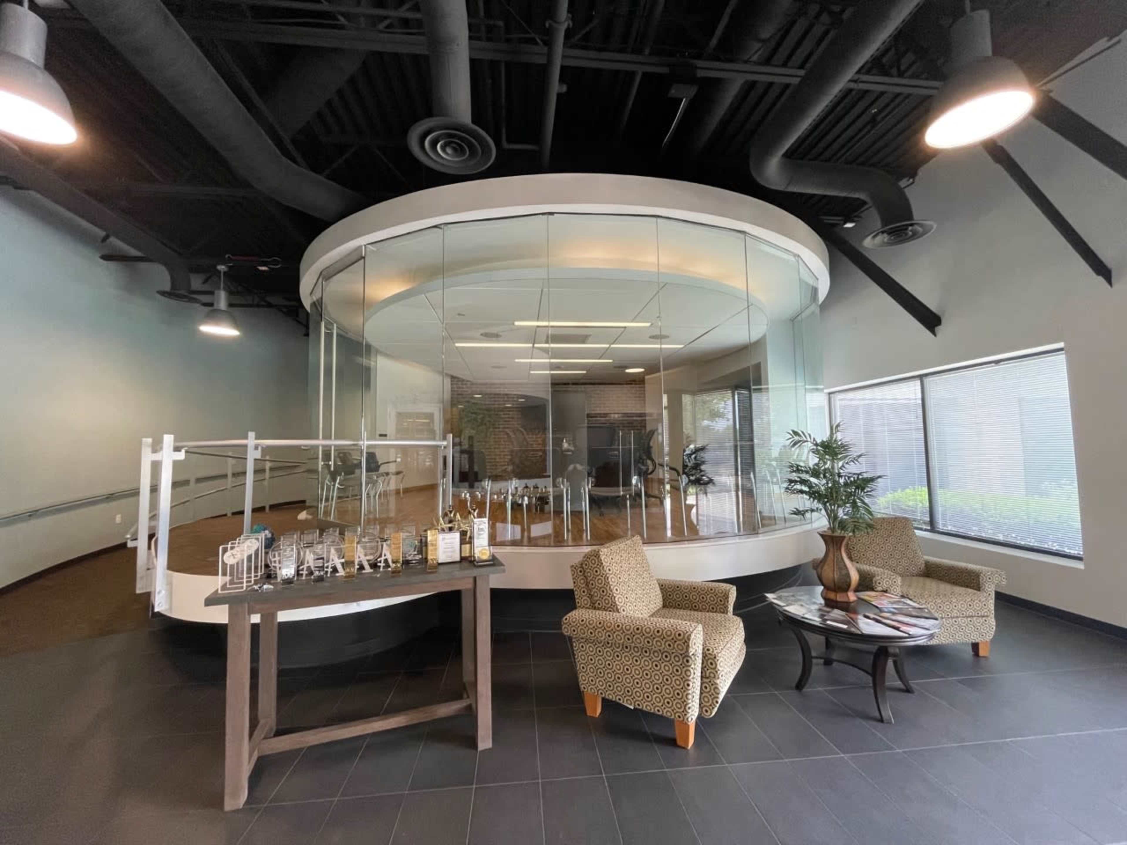 A modern lobby area featuring a circular glass meeting room, two patterned chairs, a coffee table, and a display table with awards.