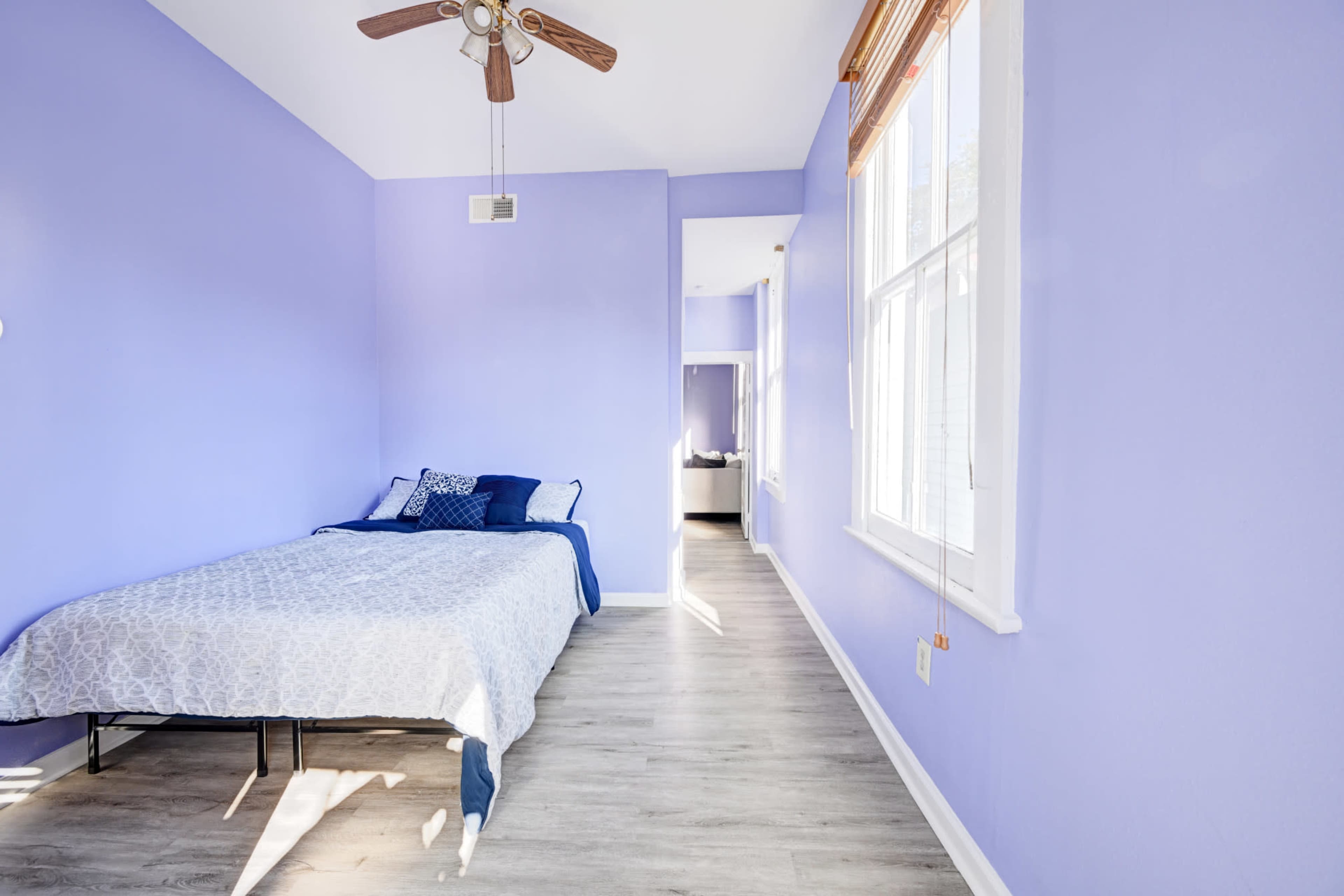 A bedroom features a bed with blue accents, light purple walls, and large windows allowing natural light to flow in.