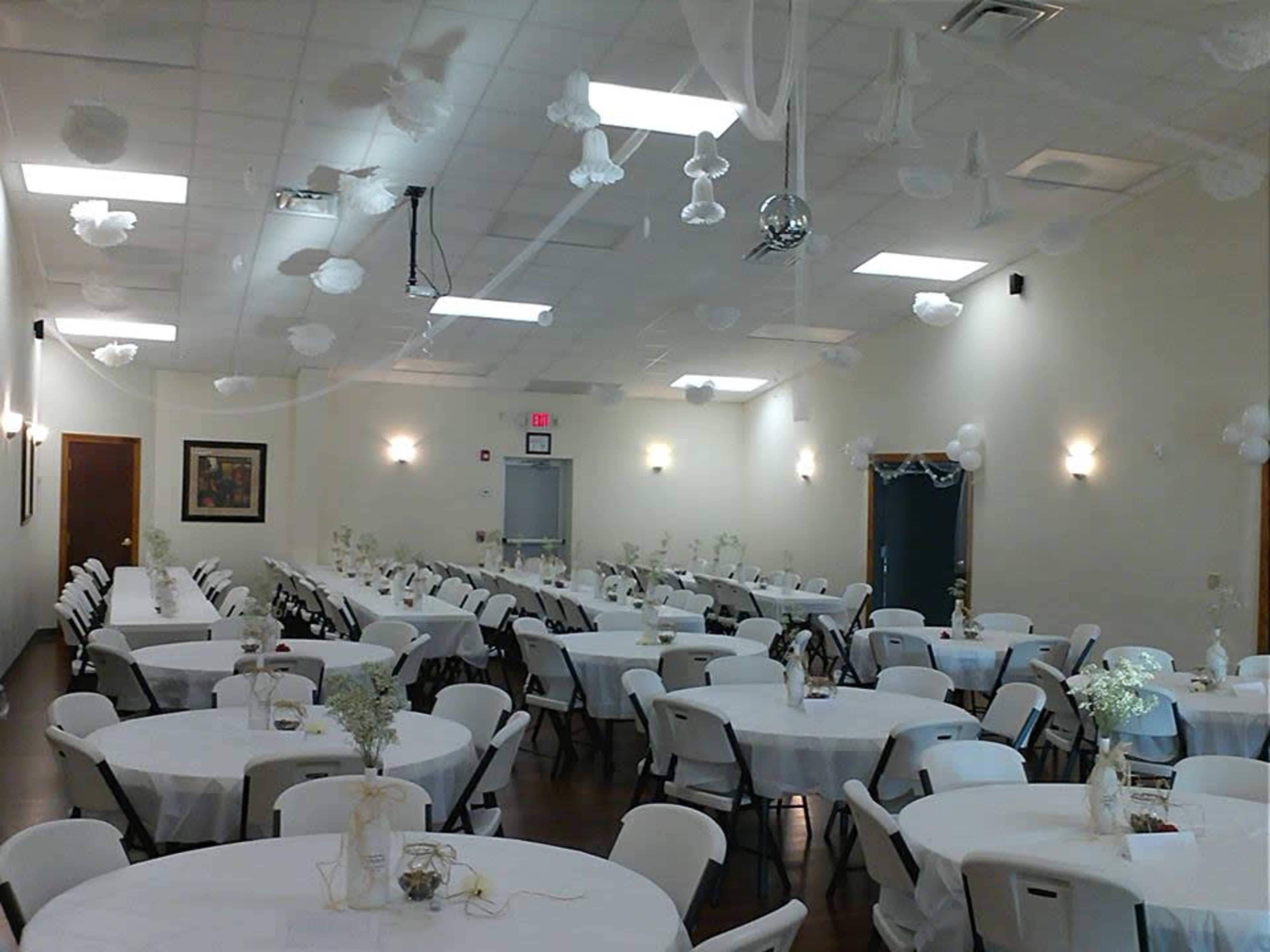 A spacious banquet hall featuring round tables covered with white tablecloths and decorated with floral arrangements, with a disco ball and white decorations hanging from the ceiling.