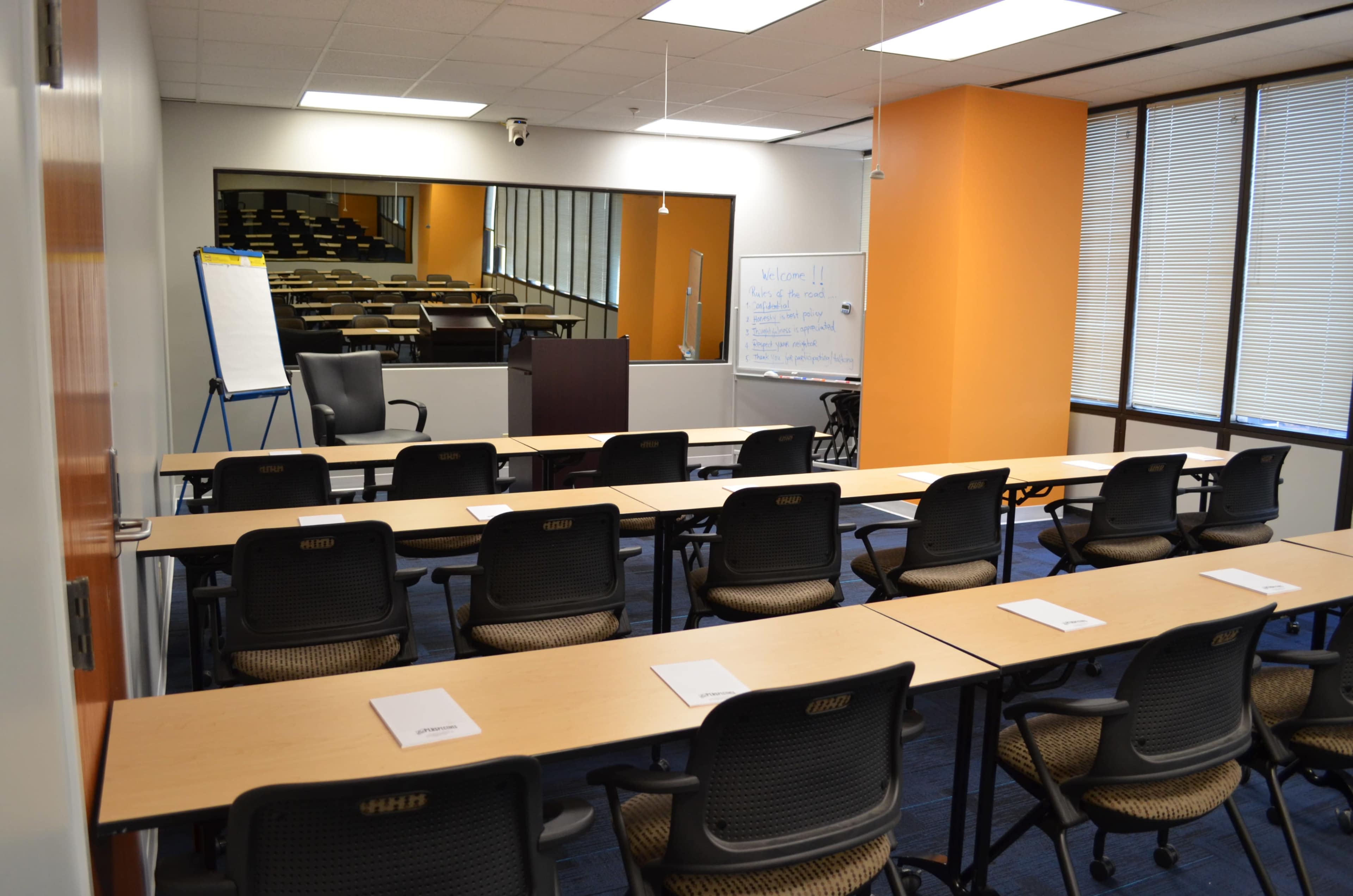 The image shows a conference room set up with rows of chairs facing a podium, whiteboard, and presentation area, with a large mirror along one wall.