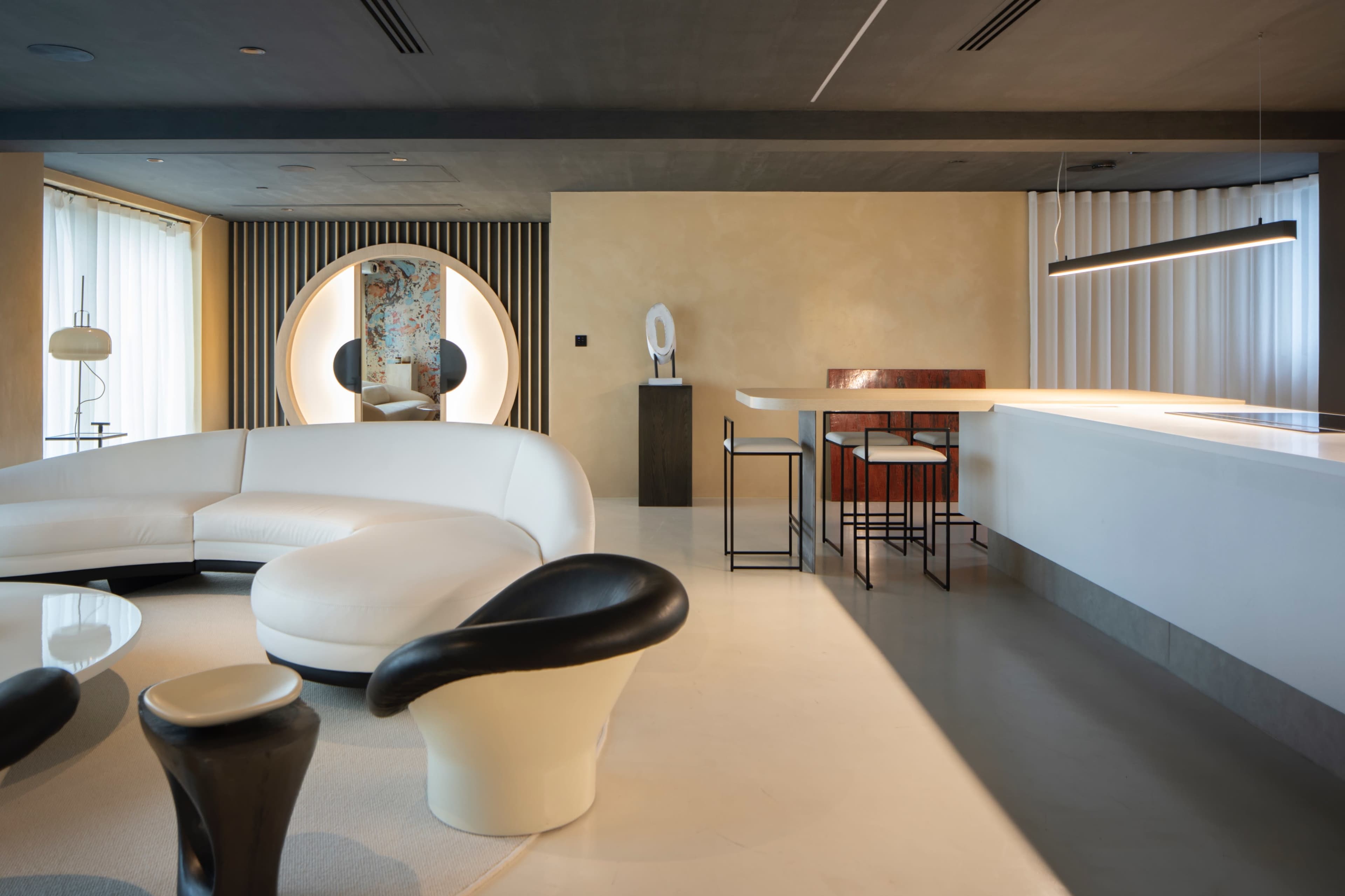 A modern interior space featuring a white curved sofa, a round mirror, a minimalist bar area with high stools, and a light-colored floor.