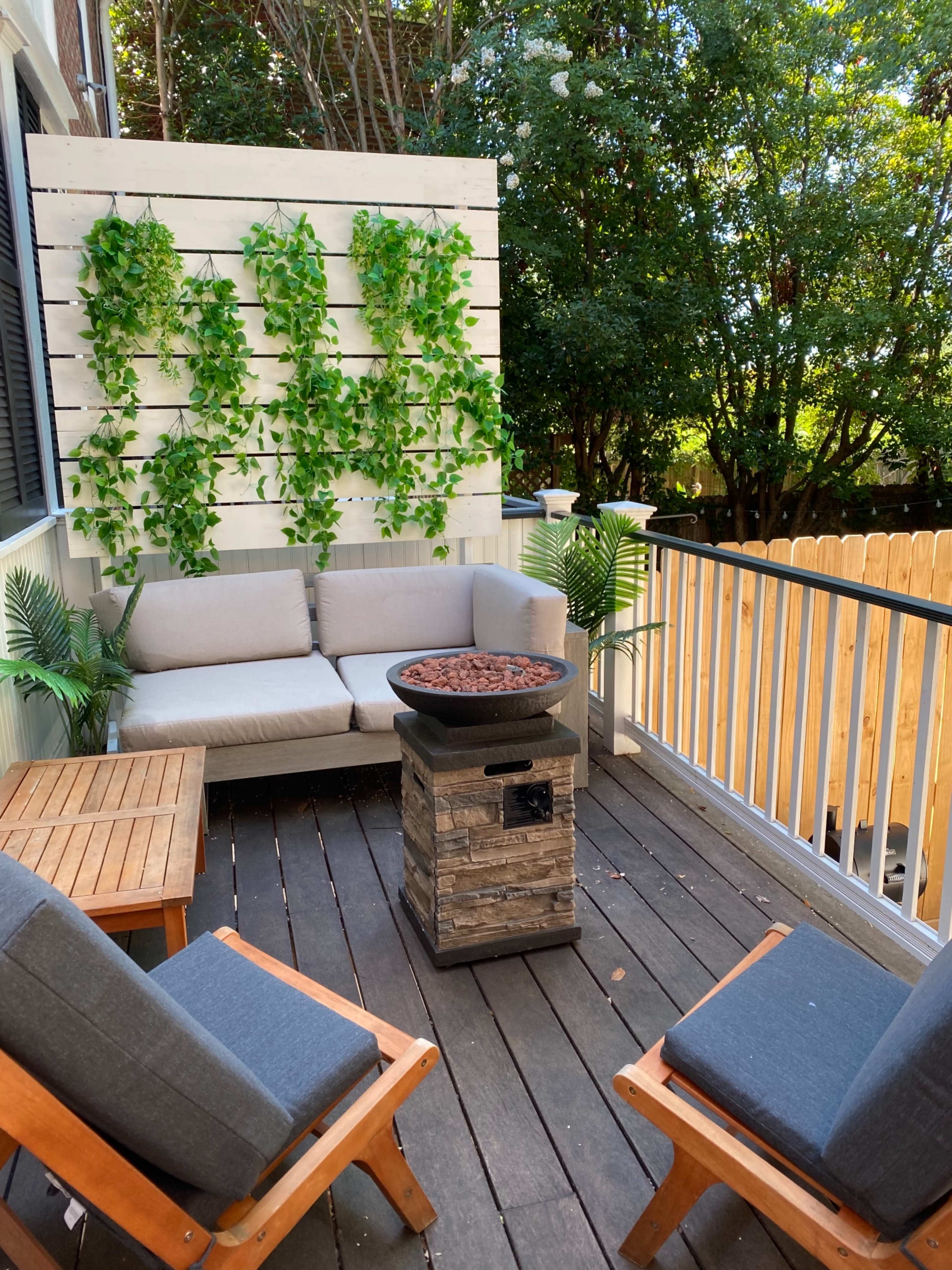 A cozy outdoor seating area with a gray couch and two wooden chairs arranged around a stone fire pit, surrounded by decorative greenery and wooden fencing.