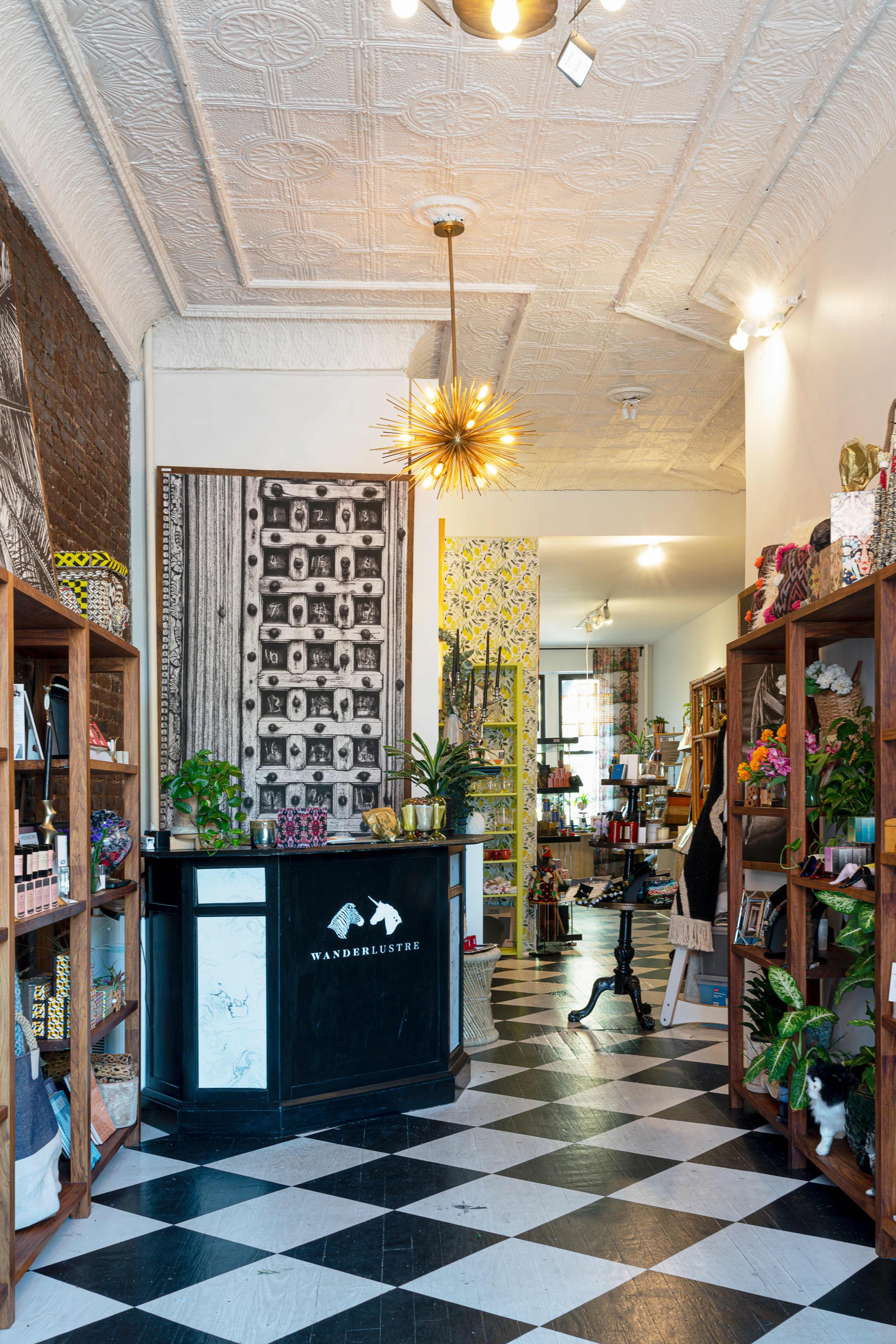 The interior of a shop features a checkered floor, wooden shelves filled with various items, and a counter displaying the name "WANDERLUSTER," surrounded by decorative plants and a patterned wall.