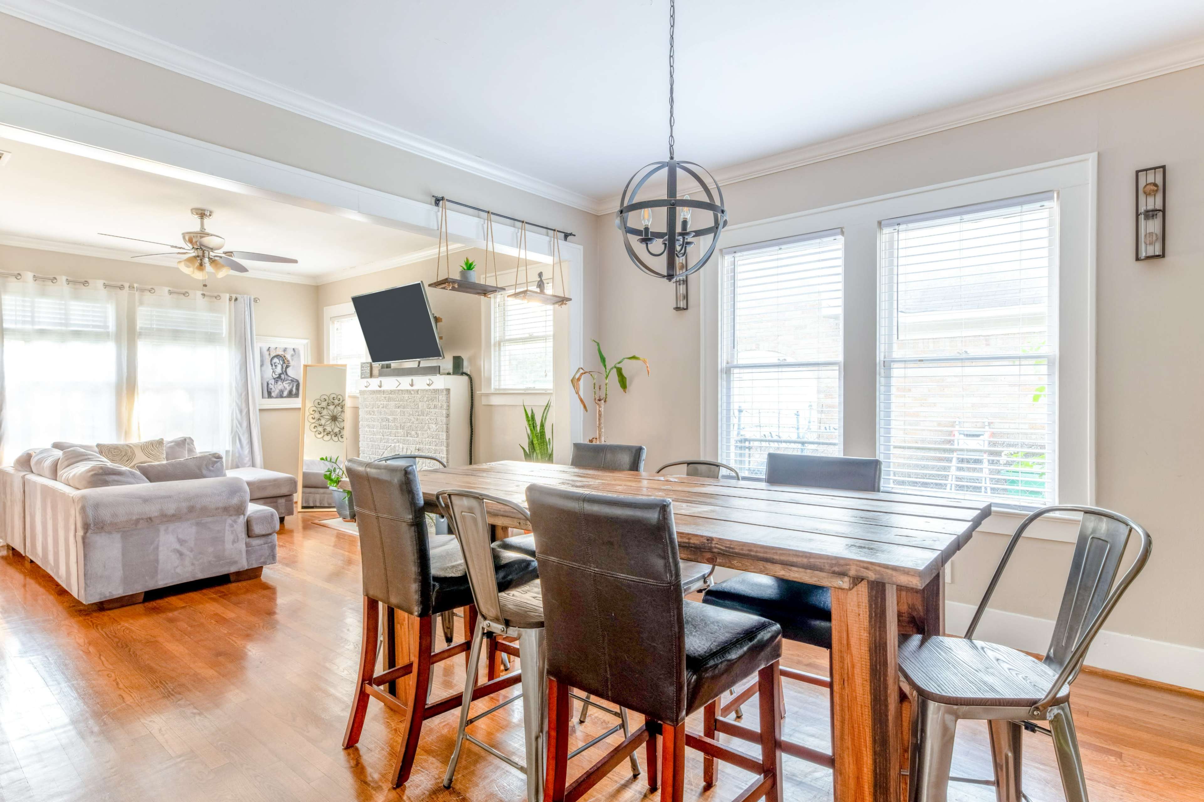 A wooden dining table with black chairs is set in a bright room featuring large windows, a comfortable couch, and a wall-mounted TV.
