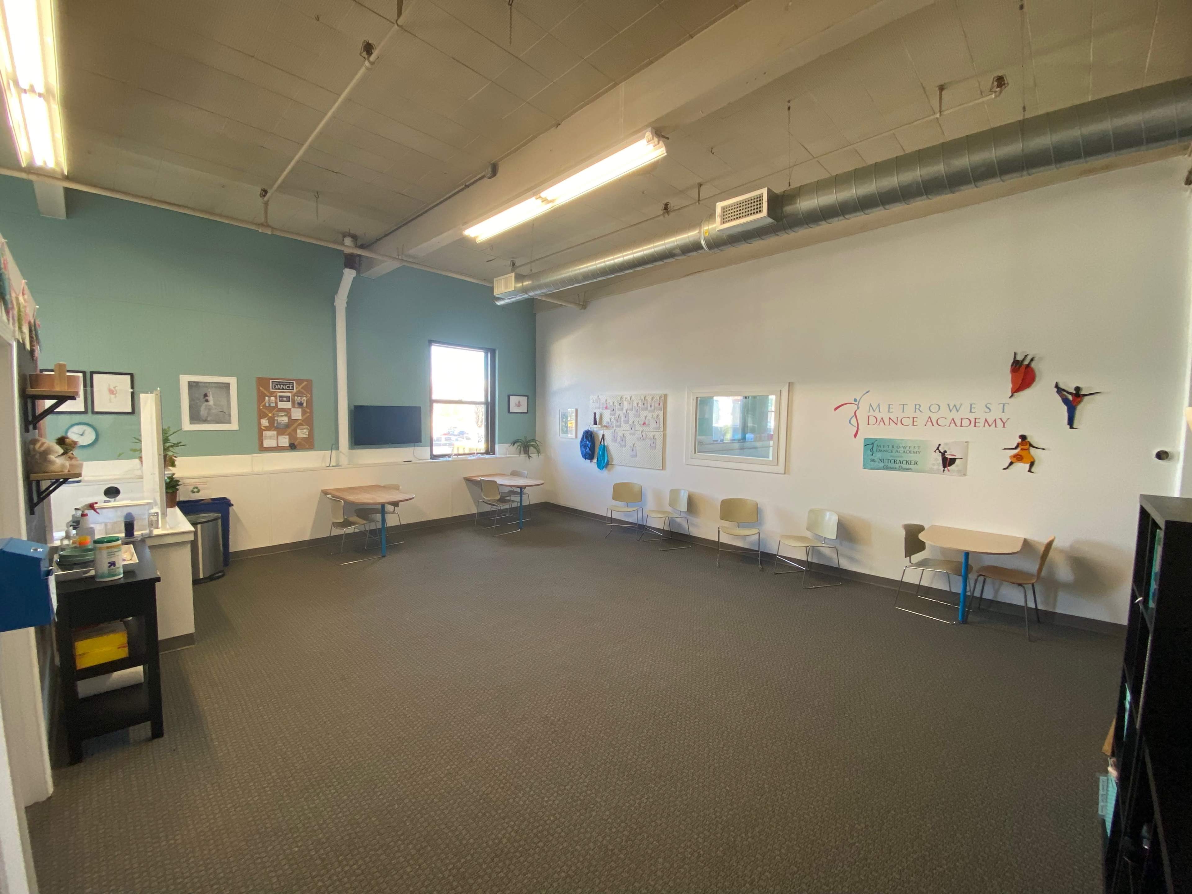 A spacious, empty classroom with several tables and chairs arranged along the walls, featuring a bulletin board and a wall adorned with dance-themed decorations.