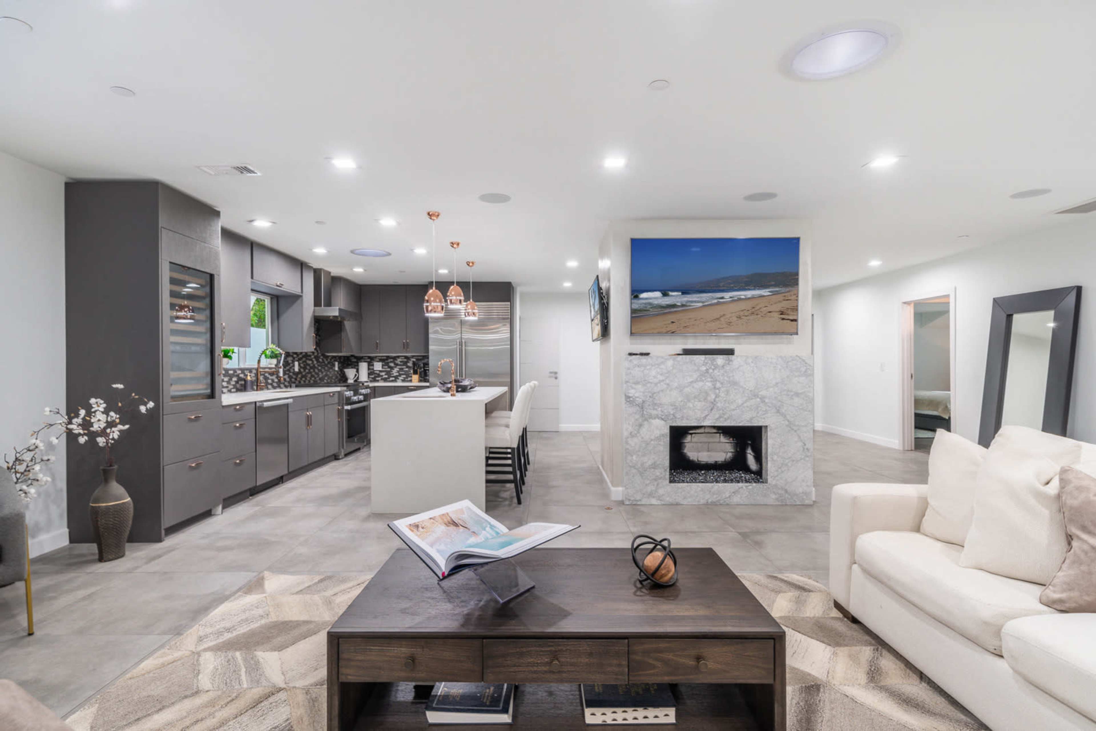 The image shows a modern kitchen and living room area with gray cabinets, a marble fireplace, and a coffee table with a book on it.