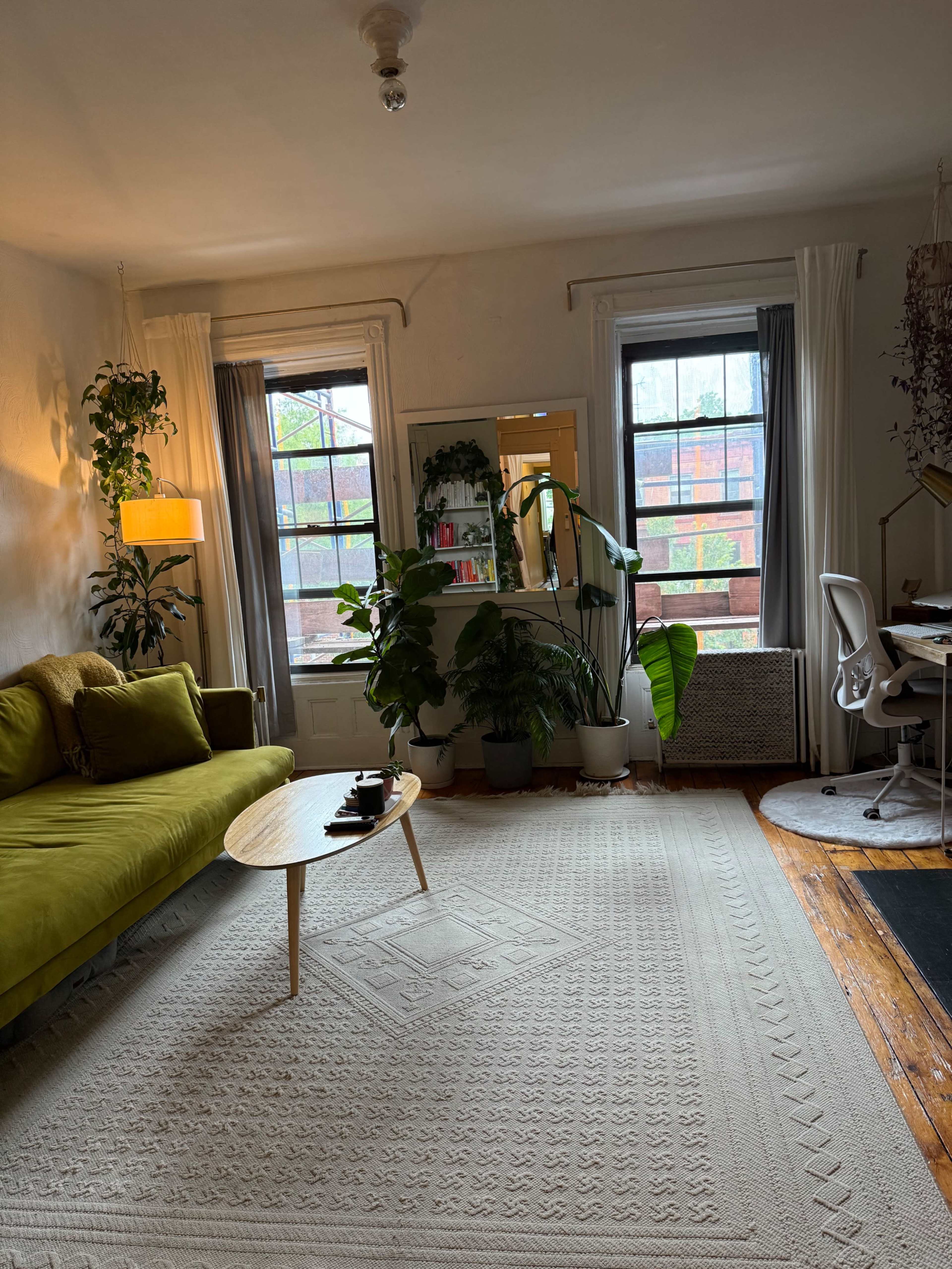 The image shows a living room with a green sofa, a round coffee table, several plants, and a workspace near a window.