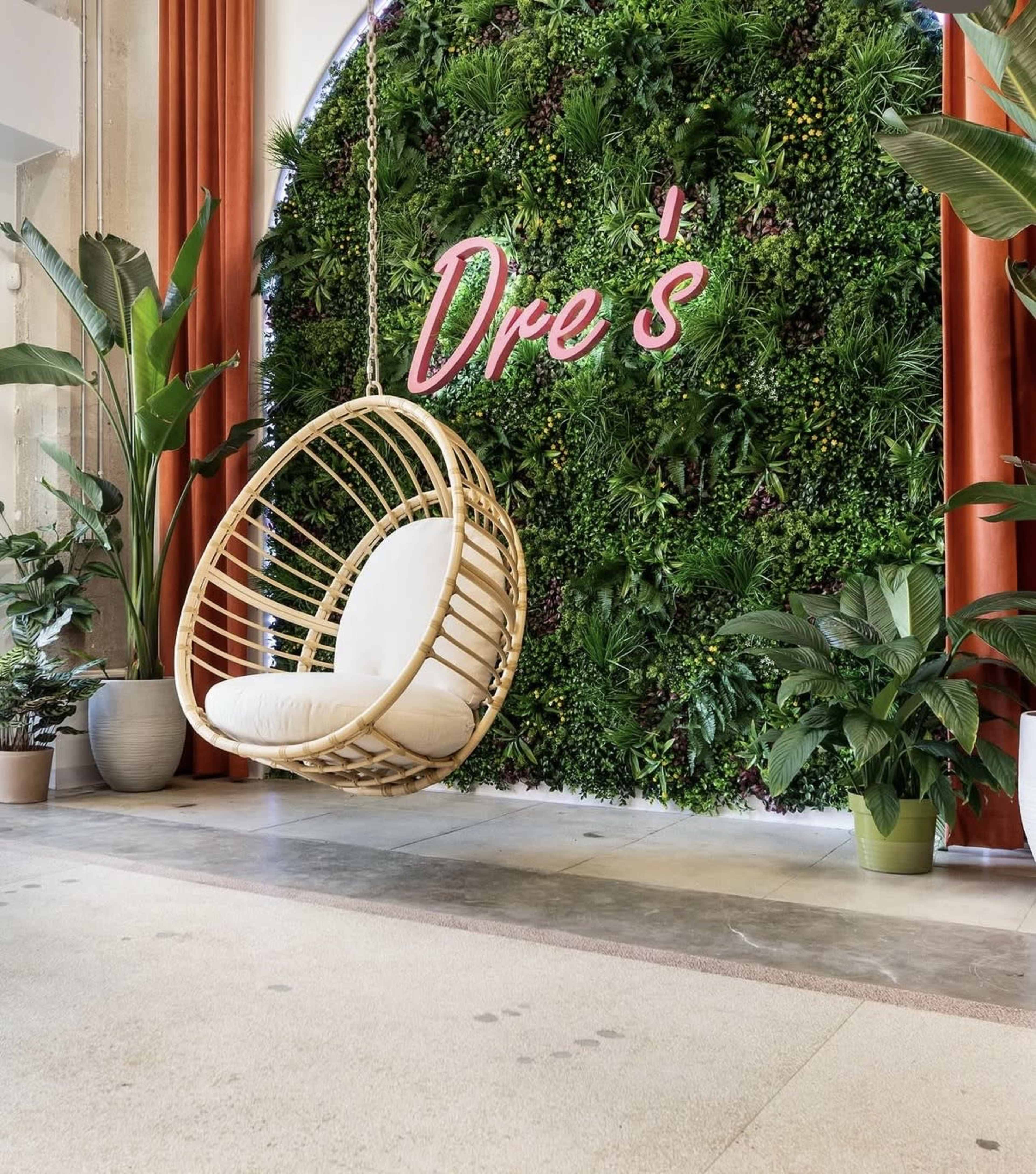 The image features a cozy indoor seating area with a hanging rattan chair in front of a vibrant green wall adorned with plants and a neon sign that reads "Dre's."