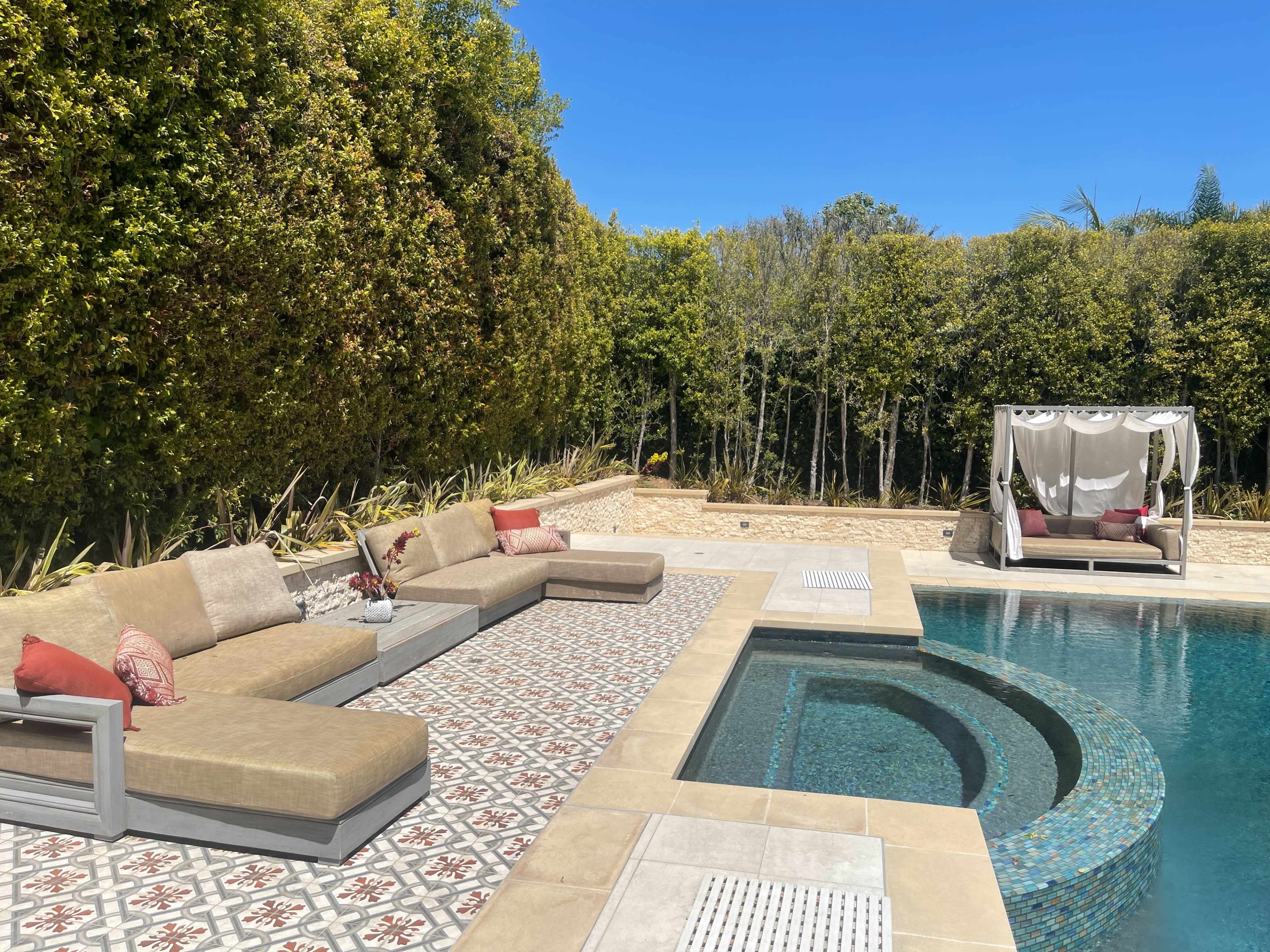 A poolside area with a large, beige sectional sofa and a shaded cabana situated near a rectangular swimming pool surrounded by greenery.