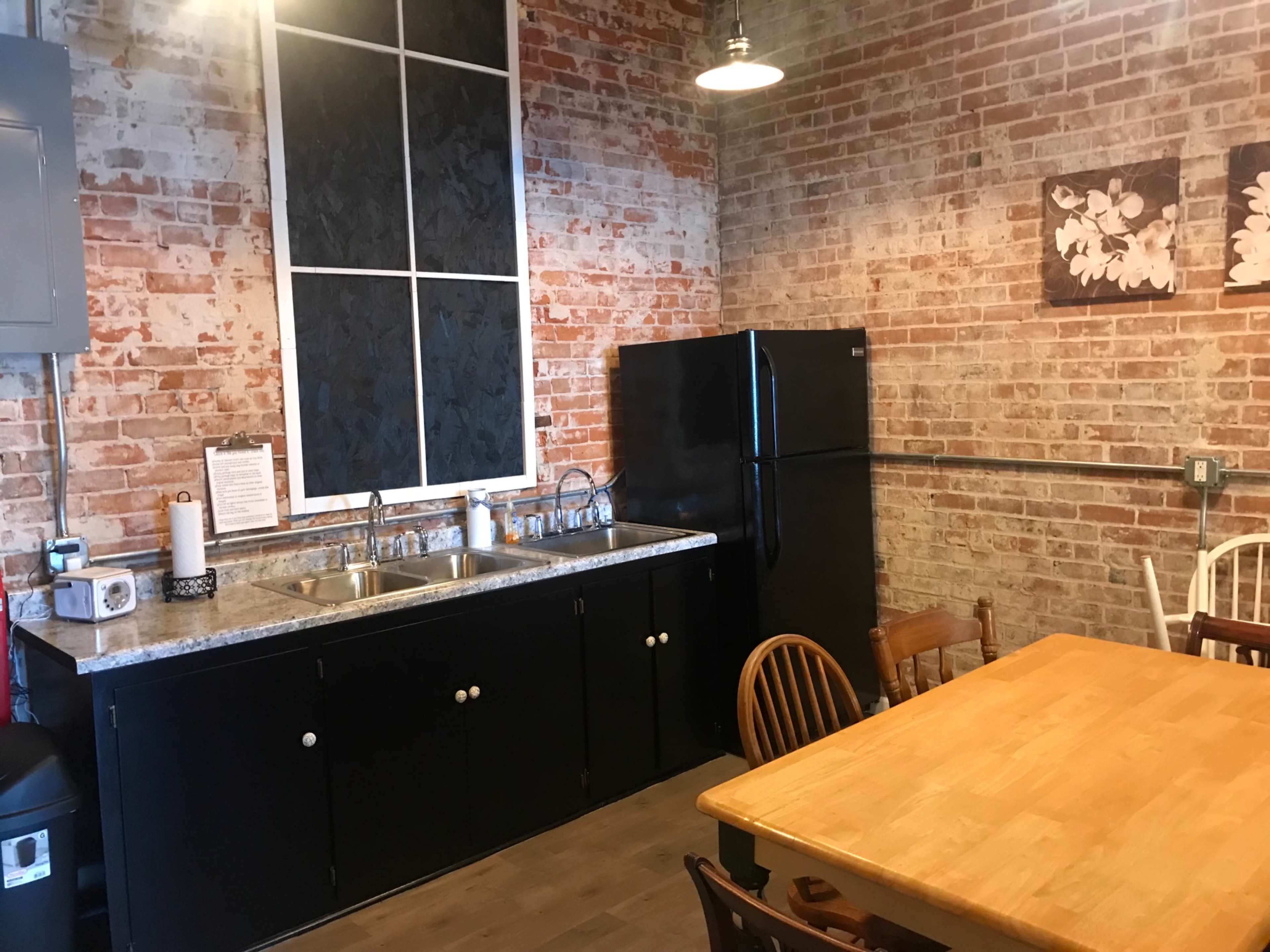 The image shows a kitchen with exposed brick walls, a black refrigerator, a double sink, and a wooden dining table surrounded by chairs.