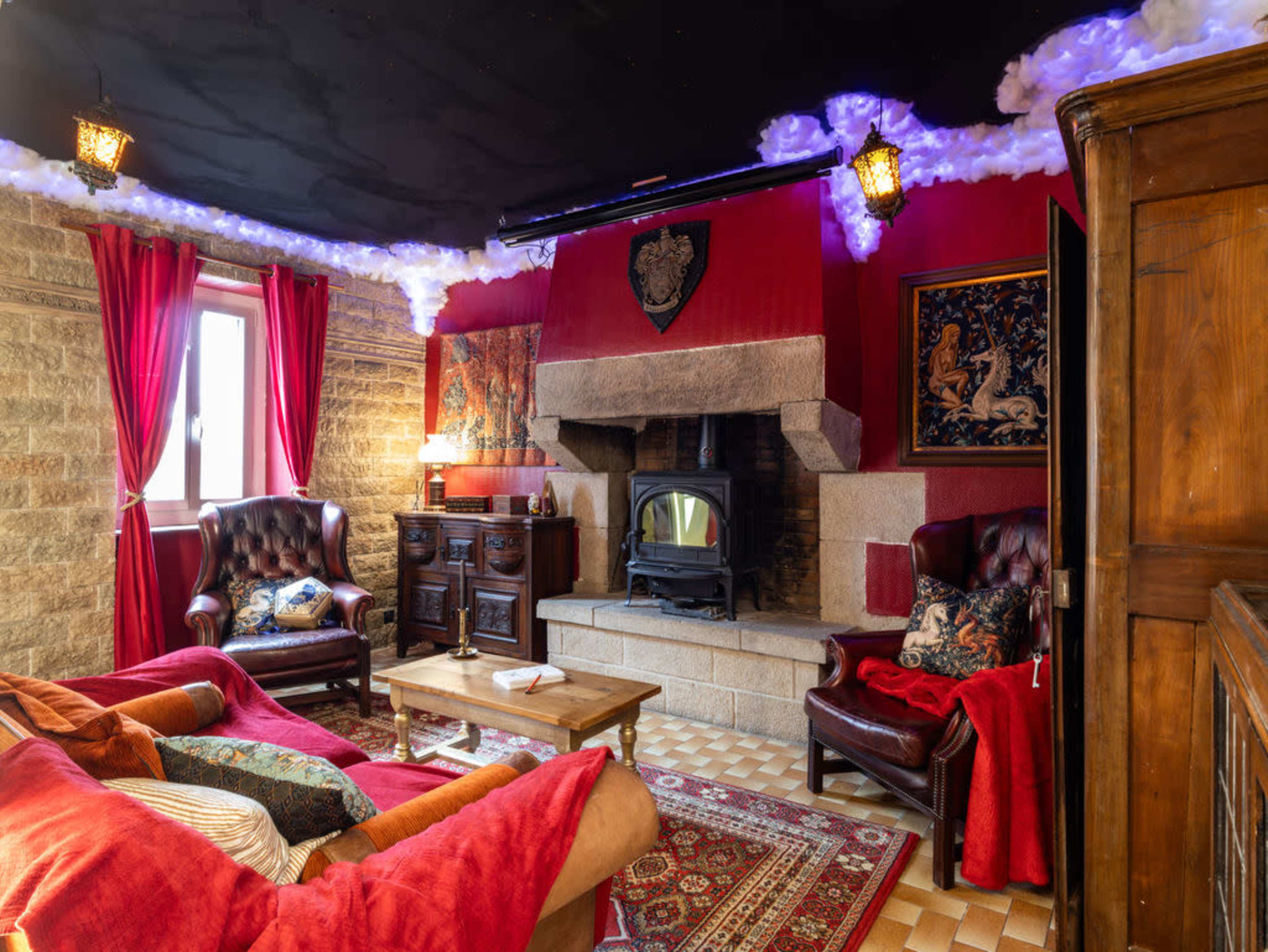 The image shows a cozy living room with a stone fireplace, red walls, and traditional furniture, including leather armchairs and a wooden cabinet.