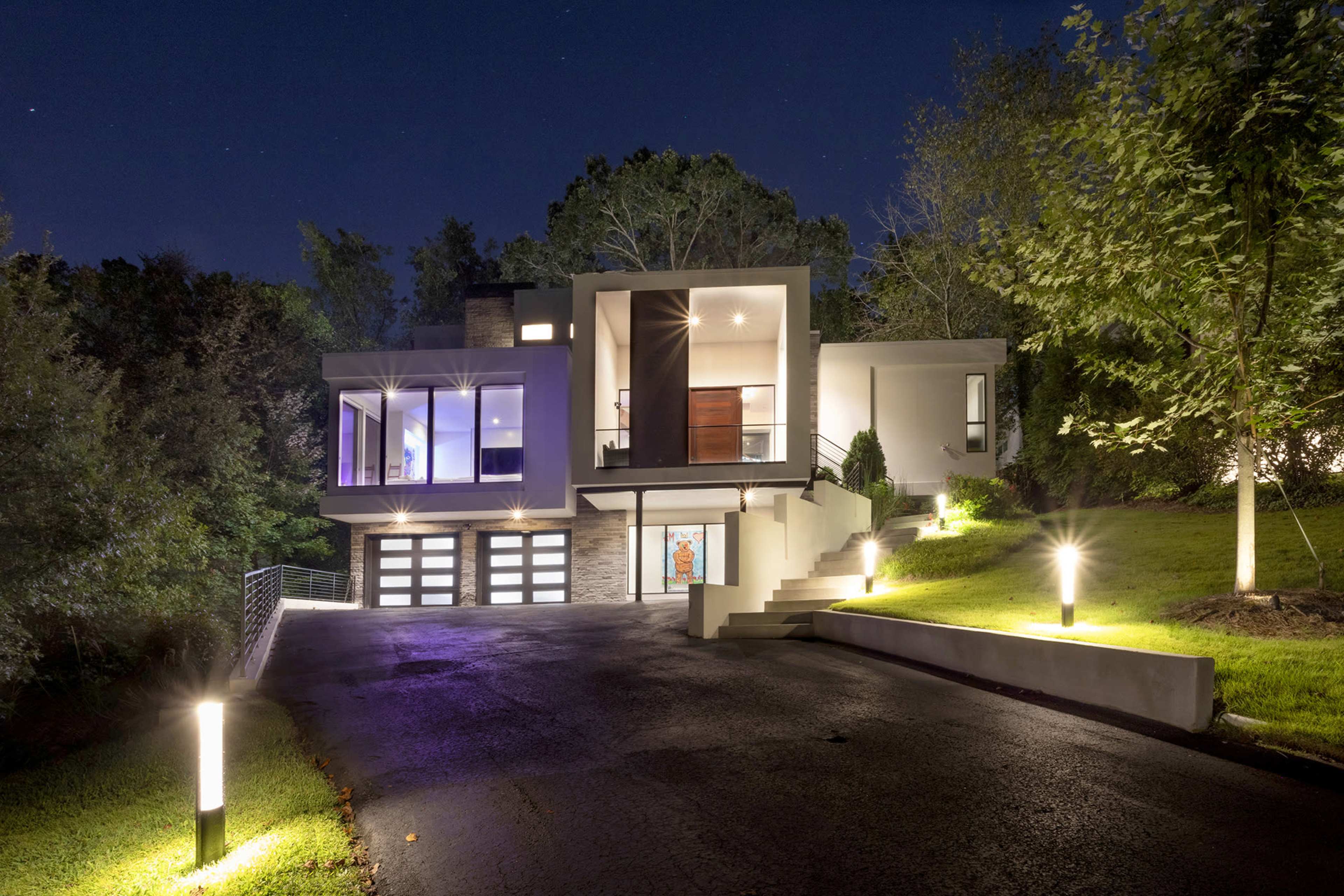 A modern house with large windows and a multi-car garage is illuminated at night, situated on a hillside surrounded by trees.