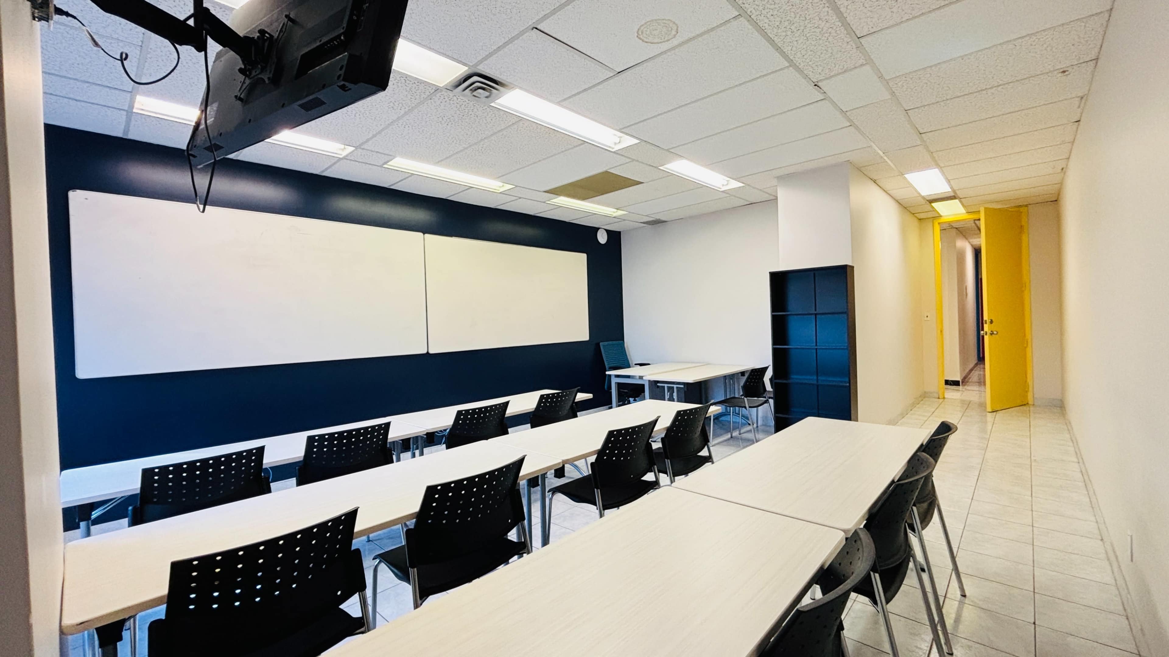 The image shows a classroom with several rows of tables and chairs, two whiteboards on the wall, and a doorway leading to another room.