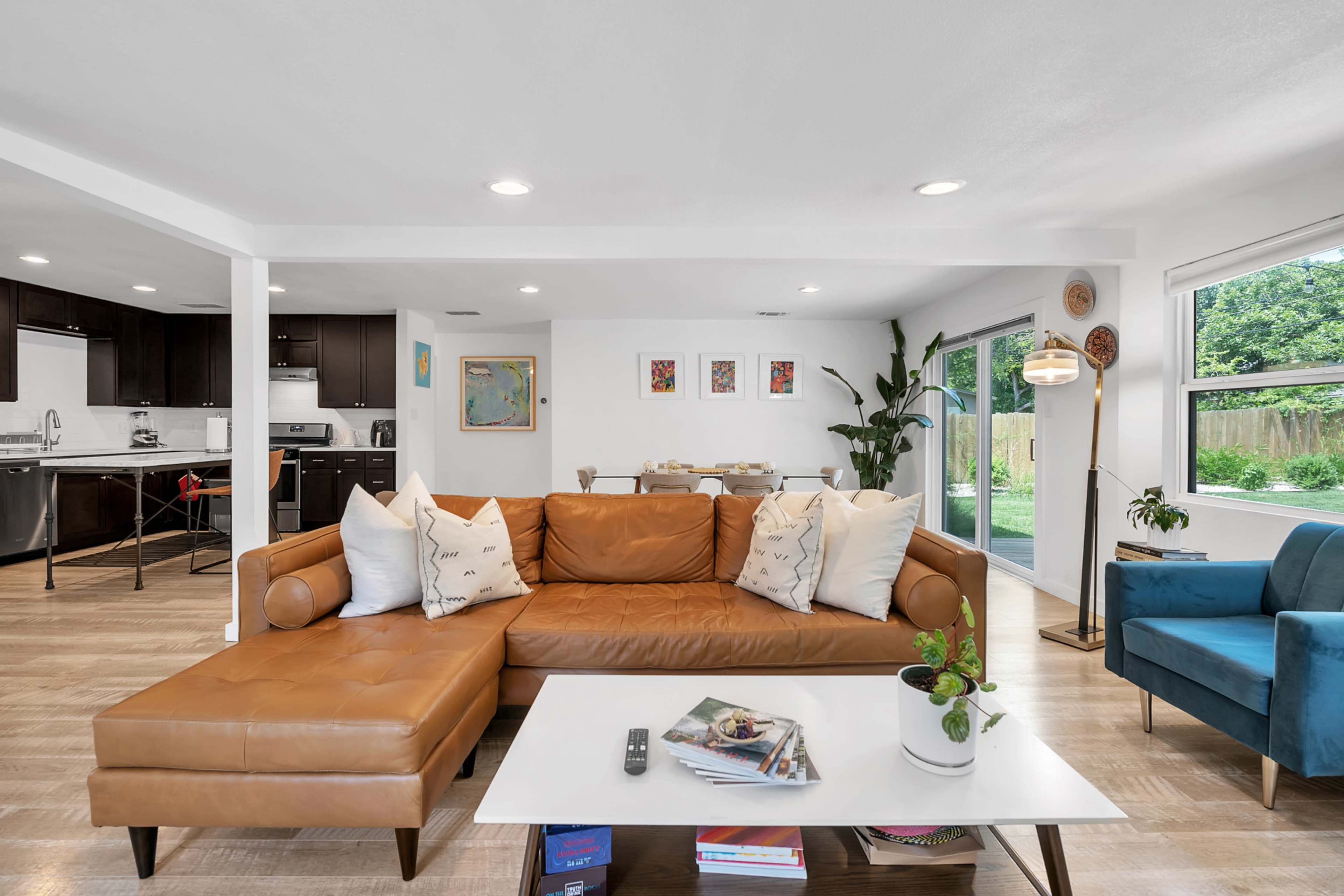 A modern living room features a brown leather sectional sofa, a white coffee table with magazines, and large windows that open to a garden area.