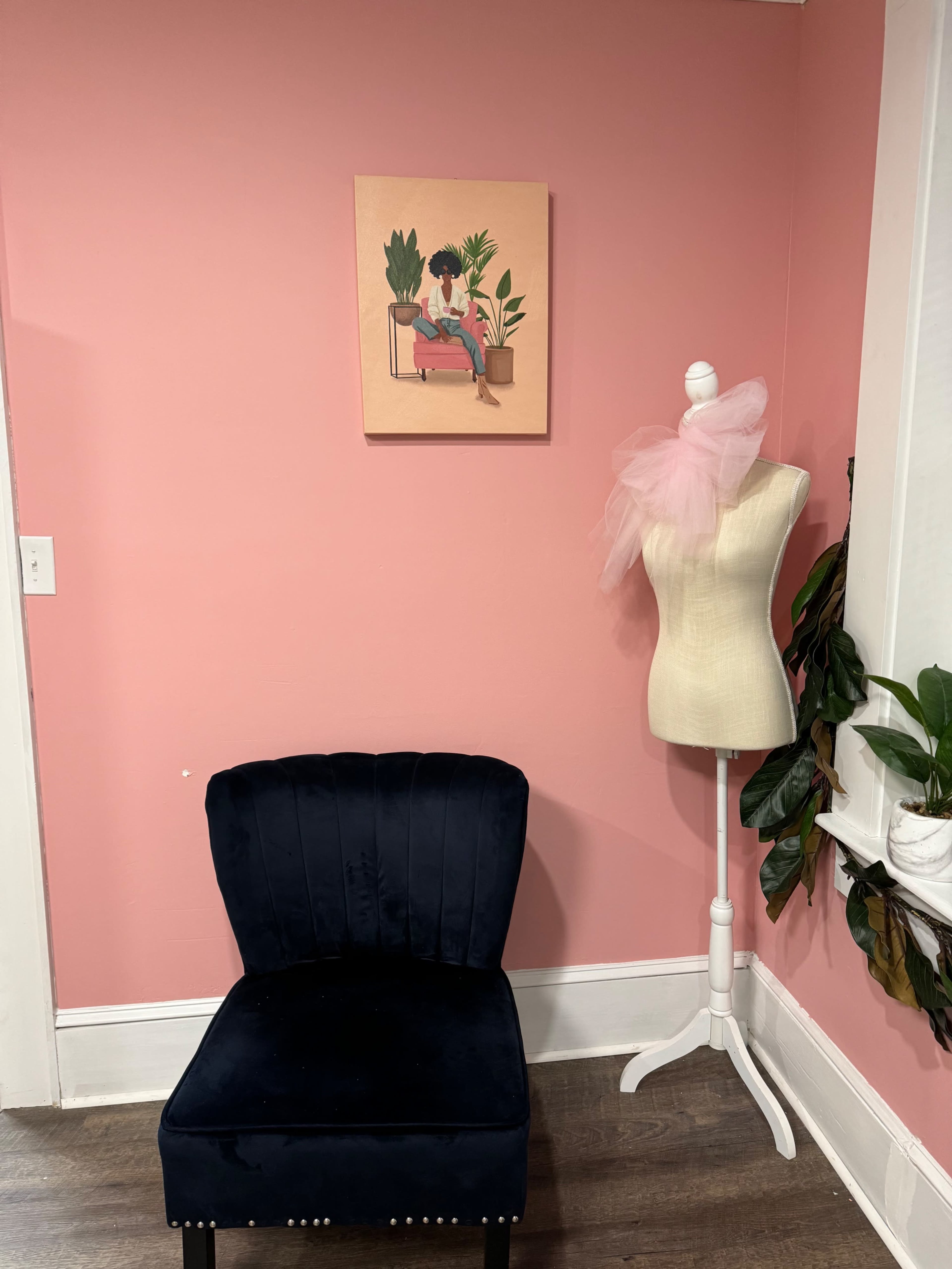The image shows a corner of a room with a pink wall, a dark blue chair, a dress form wearing a pink tulle accessory, and a framed artwork on the wall above.