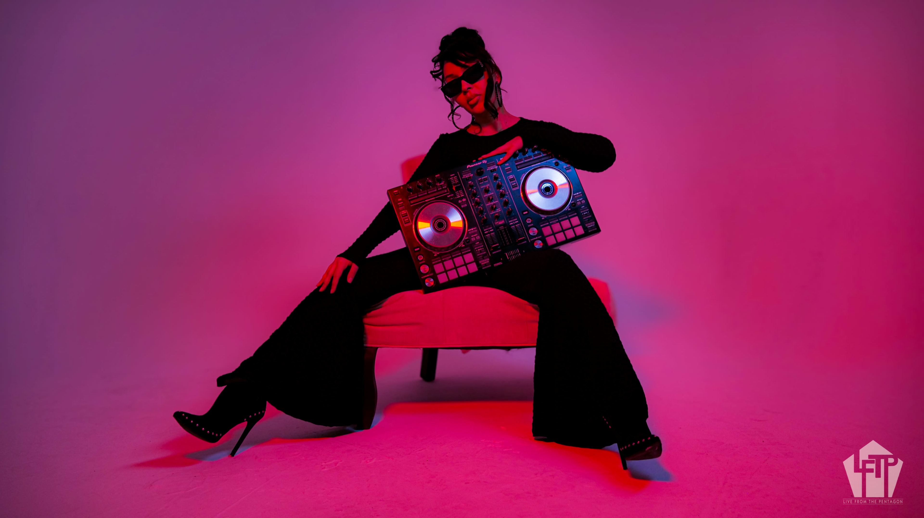 A person in a black outfit sits in a chair, holding a DJ controller, surrounded by pink and purple lighting.