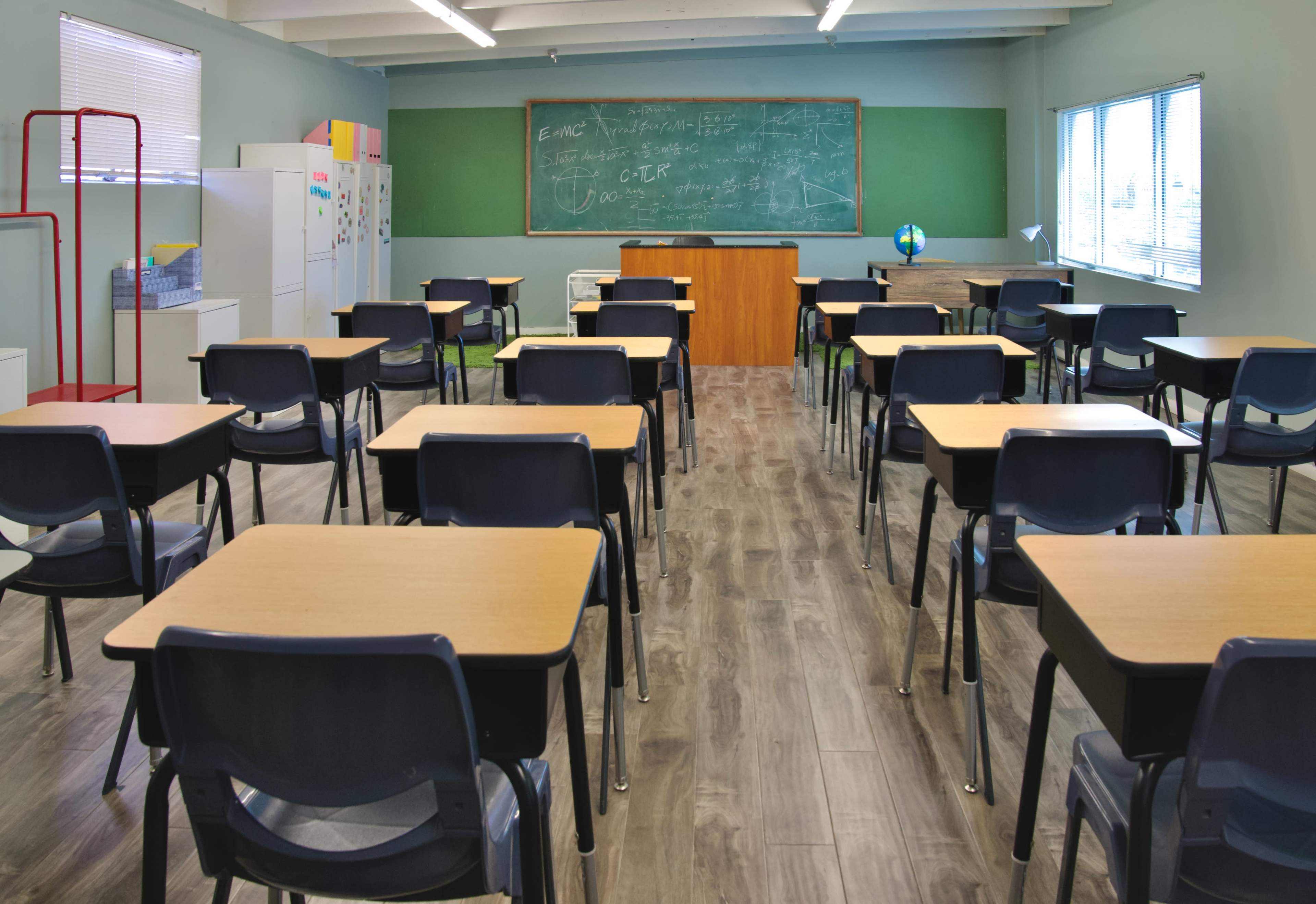 A classroom is arranged with several rows of desks facing a chalkboard and a teacher's desk at the front.