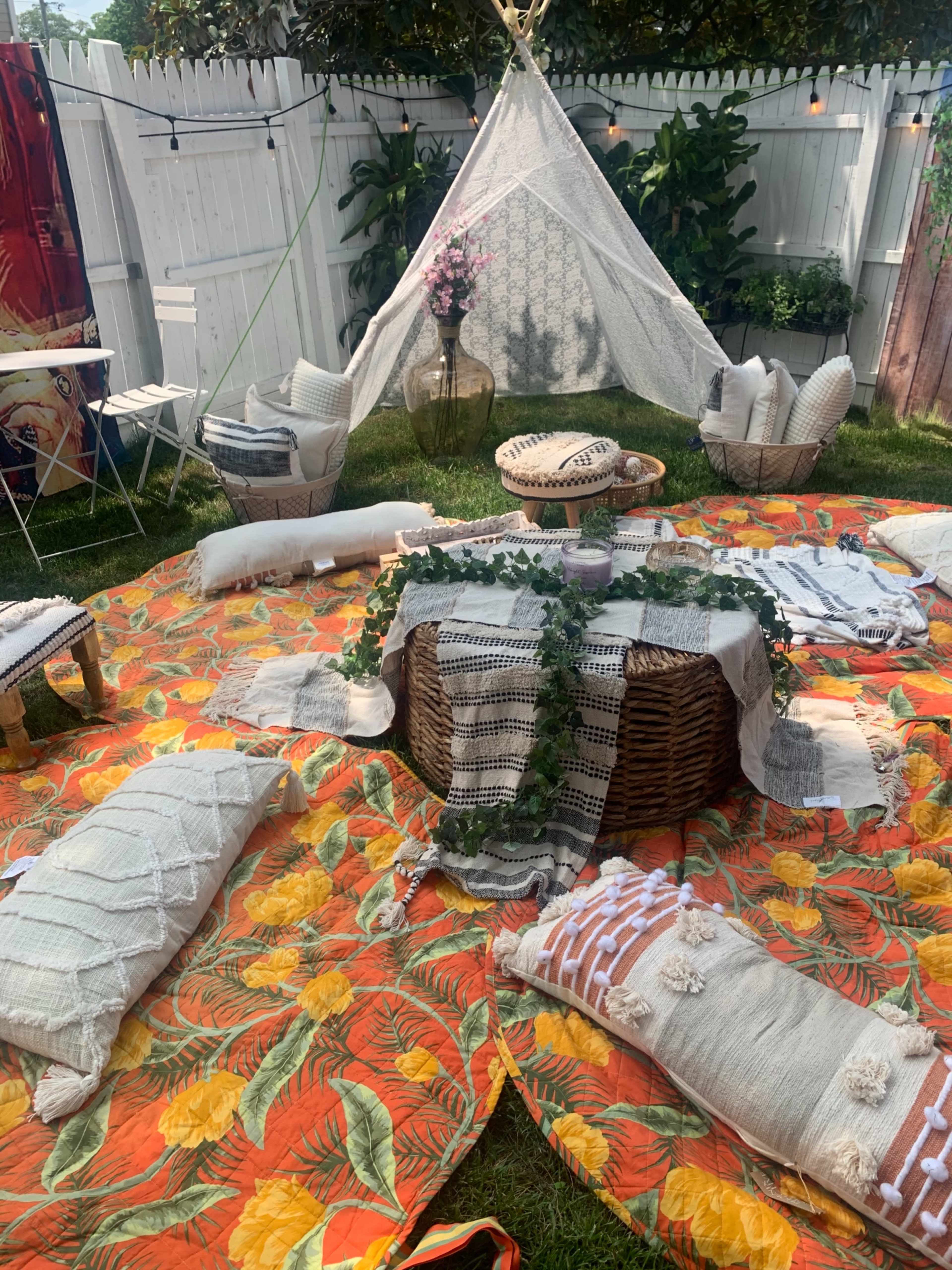 A cozy outdoor gathering space features a large circular mat surrounded by cushions, a woven table, and decorative fabrics under a tent made of lace.