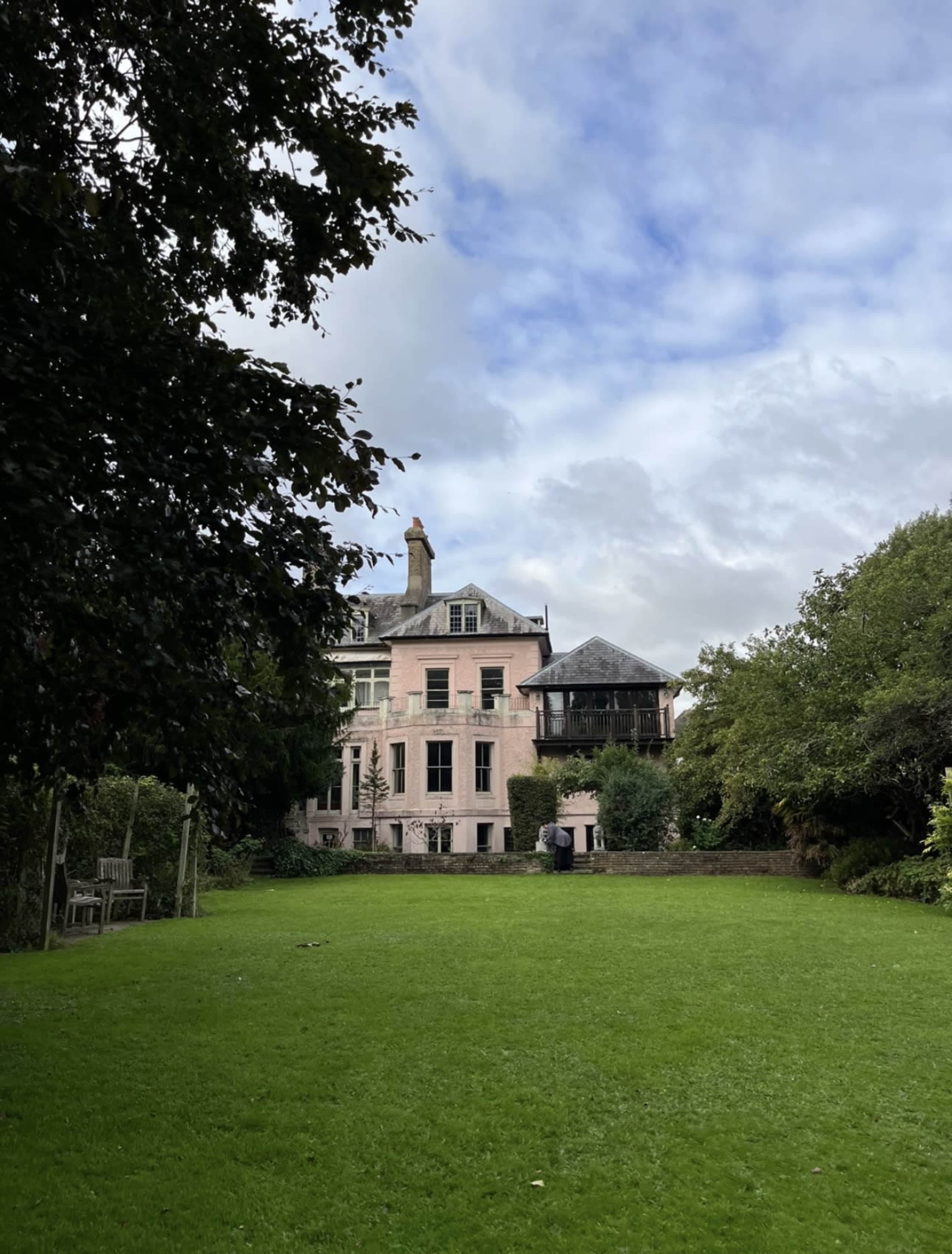 A large pink house with multiple stories is set against a cloudy sky, surrounded by a green lawn and trees.
