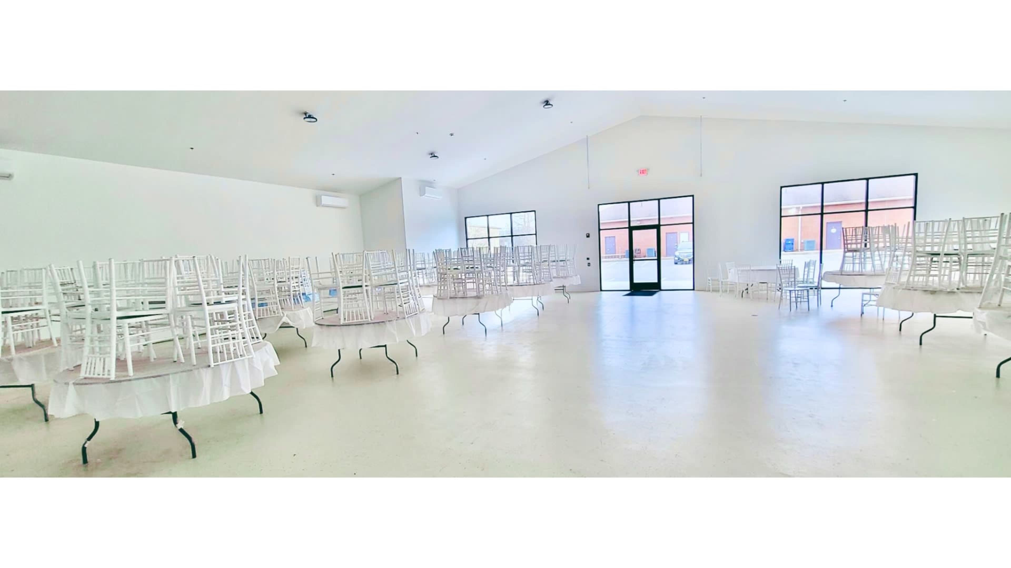 An empty indoor space features several tables with stacked white chairs and a large door leading outside.