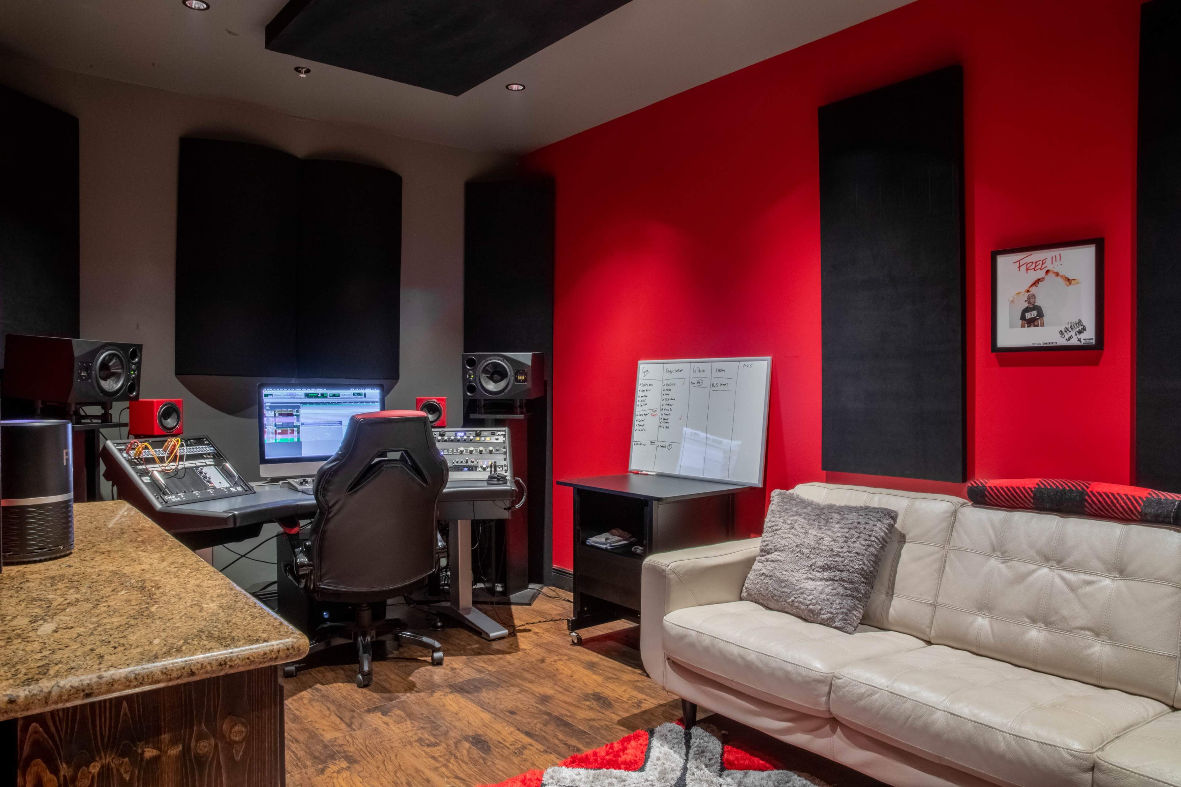A modern recording studio featuring a central workstation with speakers, a computer monitor, and a comfortable white couch against a backdrop of red and black walls.