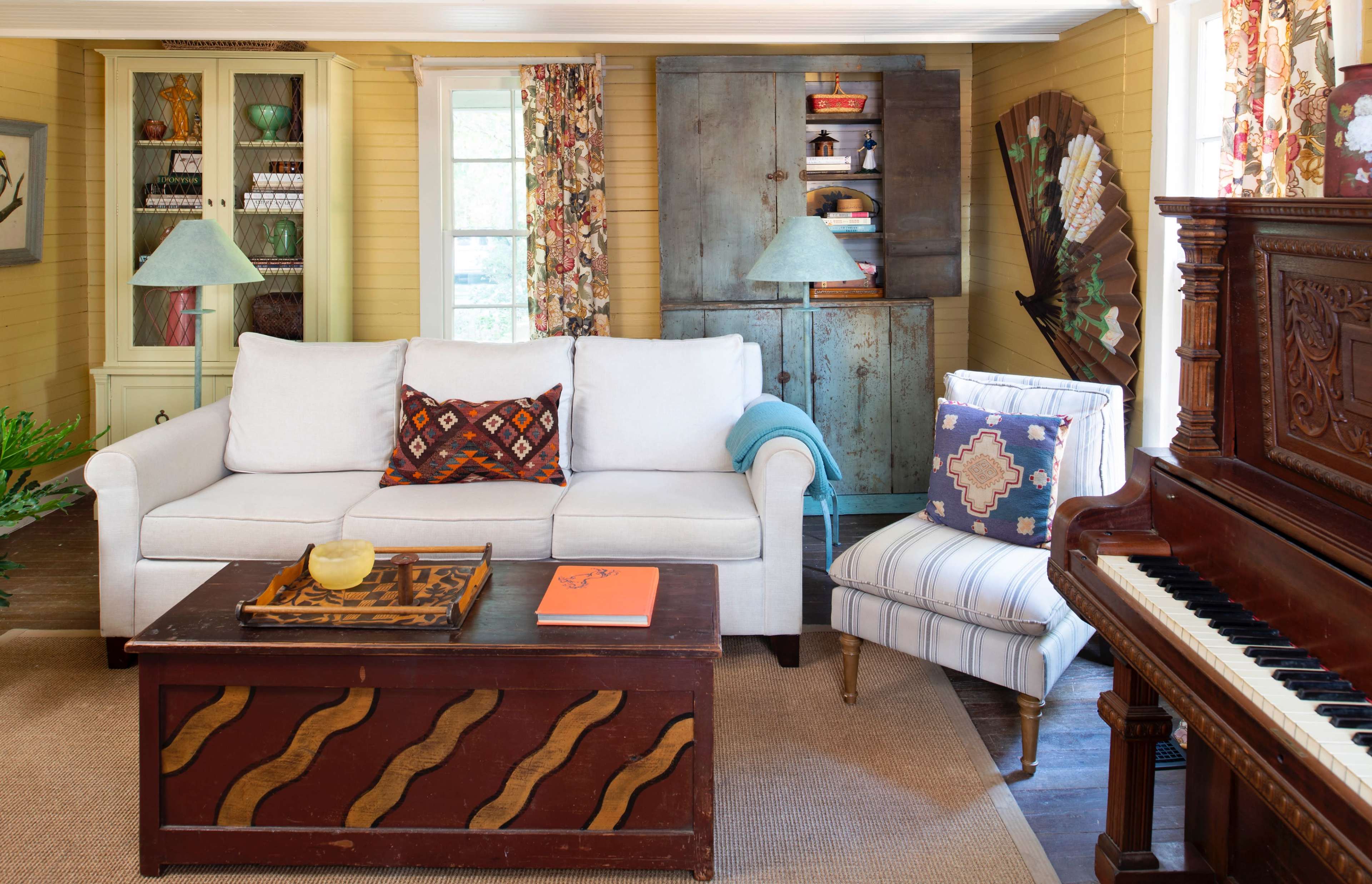 The living room features a white sofa, a wooden coffee table with wavy patterns, a striped chair, and a piano against a backdrop of yellow walls and decorative furnishings.