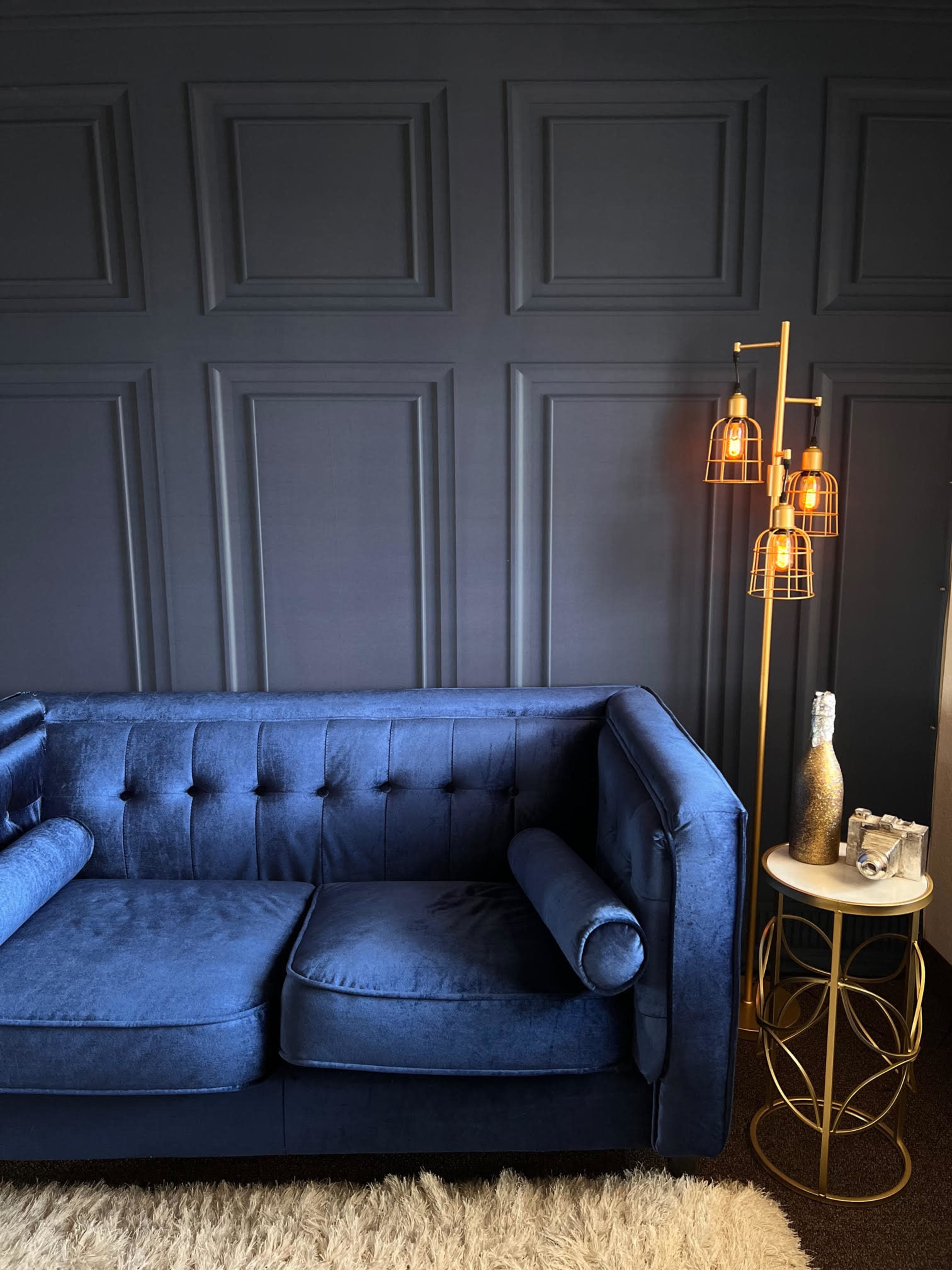 The image shows a blue velvet couch against a dark paneled wall, accompanied by a gold lamp and a circular side table.