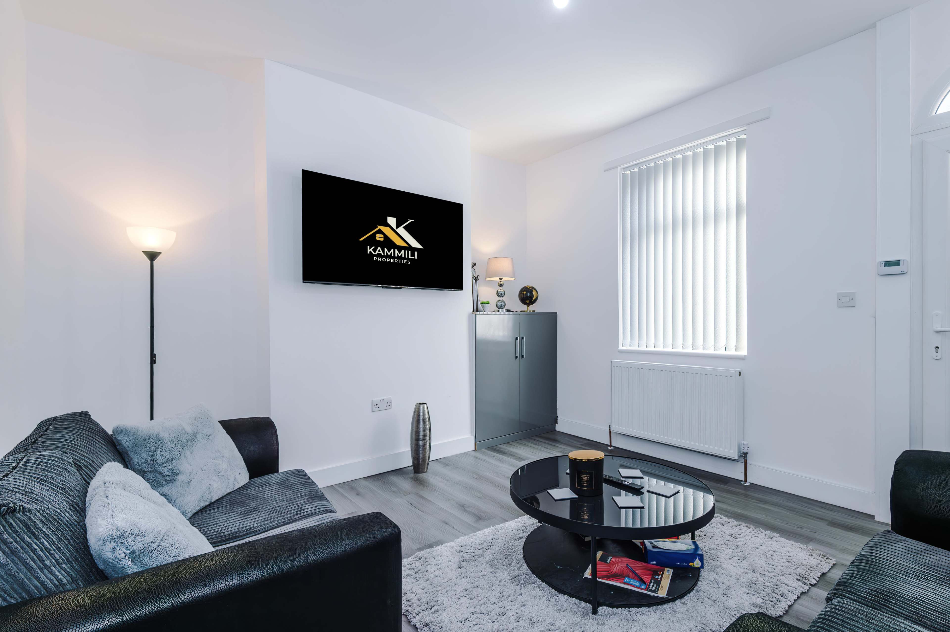A modern living room features a black sofa, a round coffee table, and a wall-mounted television displaying a logo, with a window that has vertical blinds.