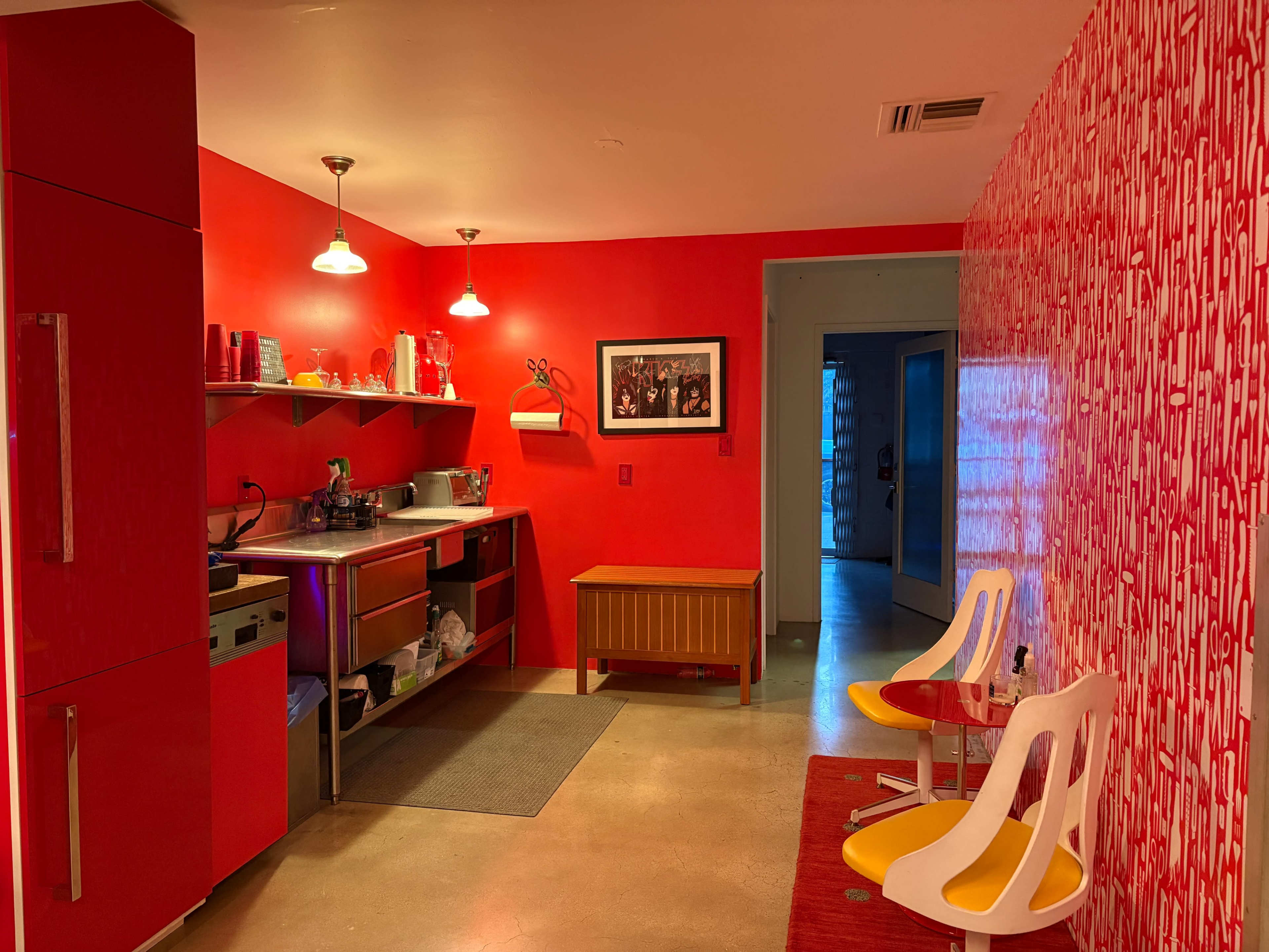 The image shows a brightly colored kitchen with red walls, a stainless steel counter, and modern furniture including white chairs.