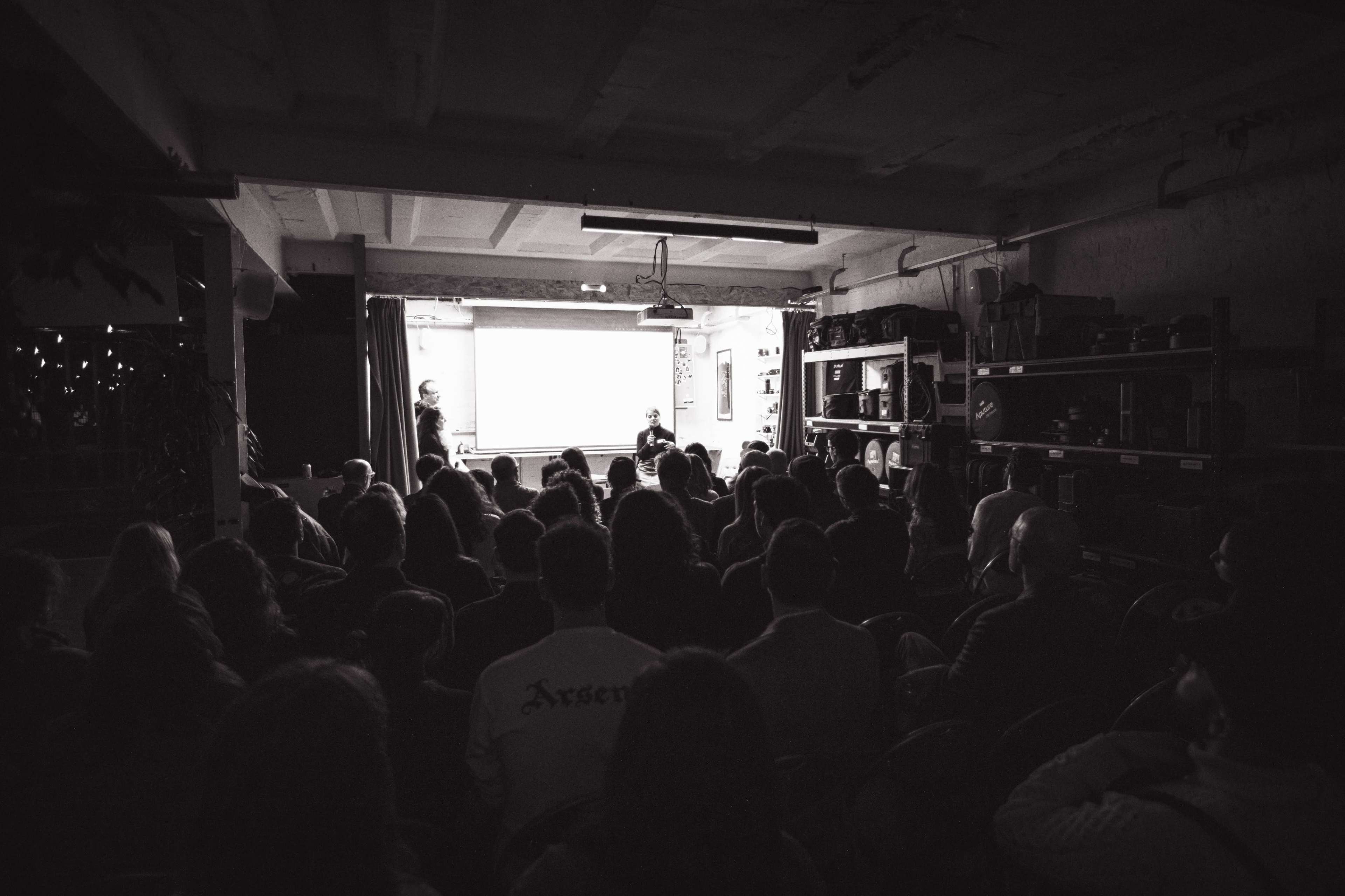 A crowd of people sits in a dimly lit room, facing a projector screen where a presentation is taking place.