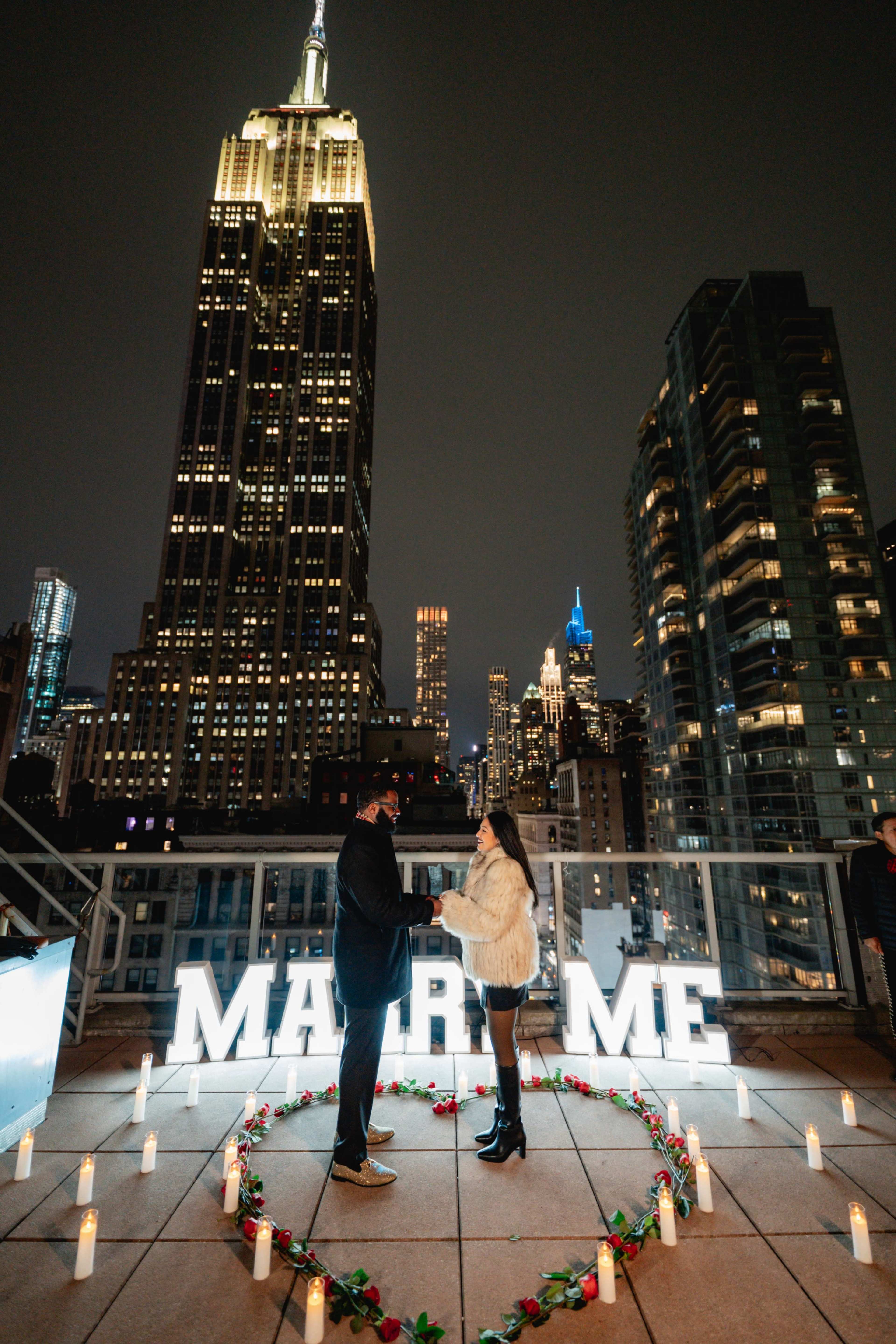 A couple stands together in front of a large "MARRY ME" sign on a rooftop with the Empire State Building illuminated in the background.