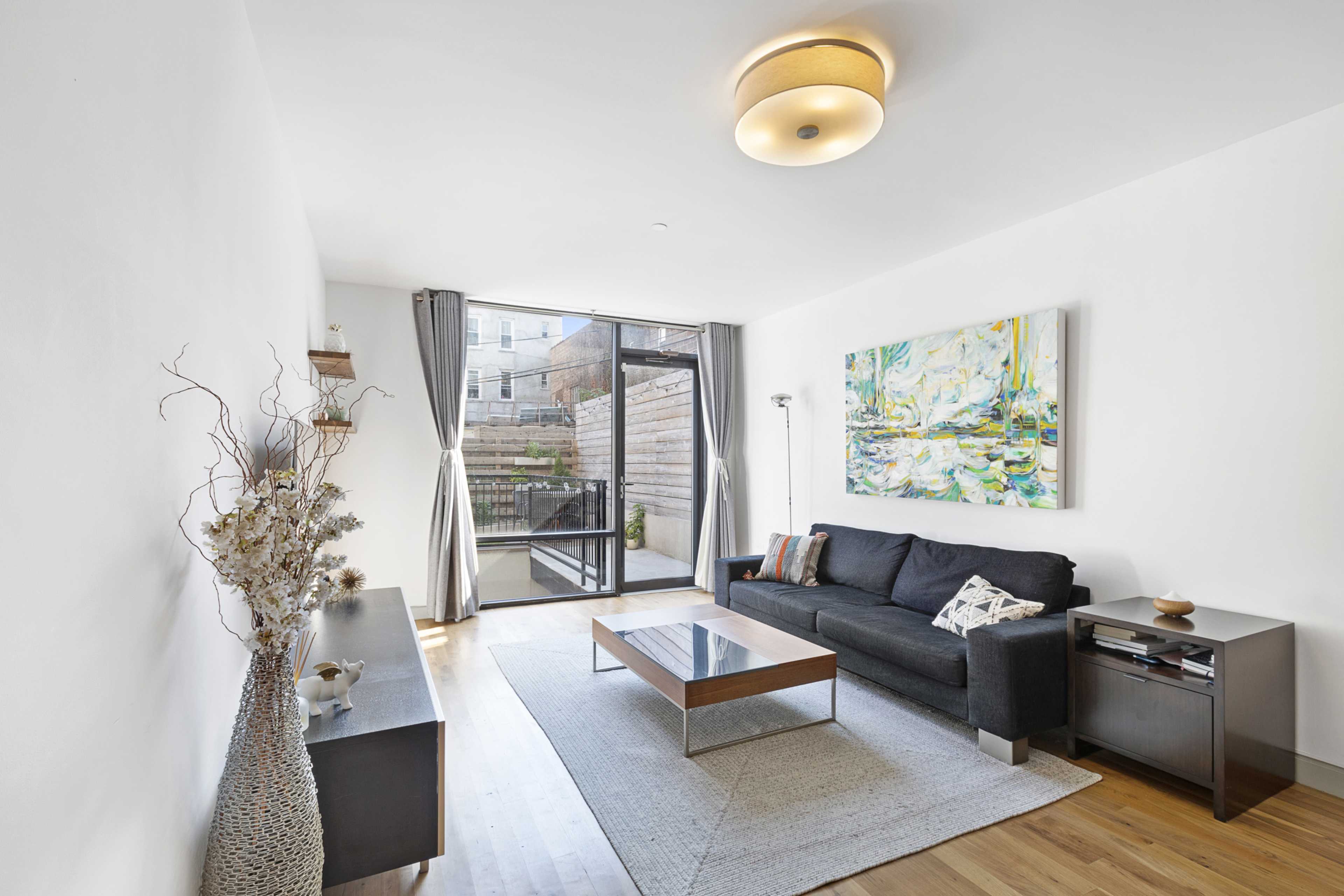 A modern living room features a dark sofa, a coffee table, and large glass doors leading to an outdoor area, with a piece of art displayed on the wall.