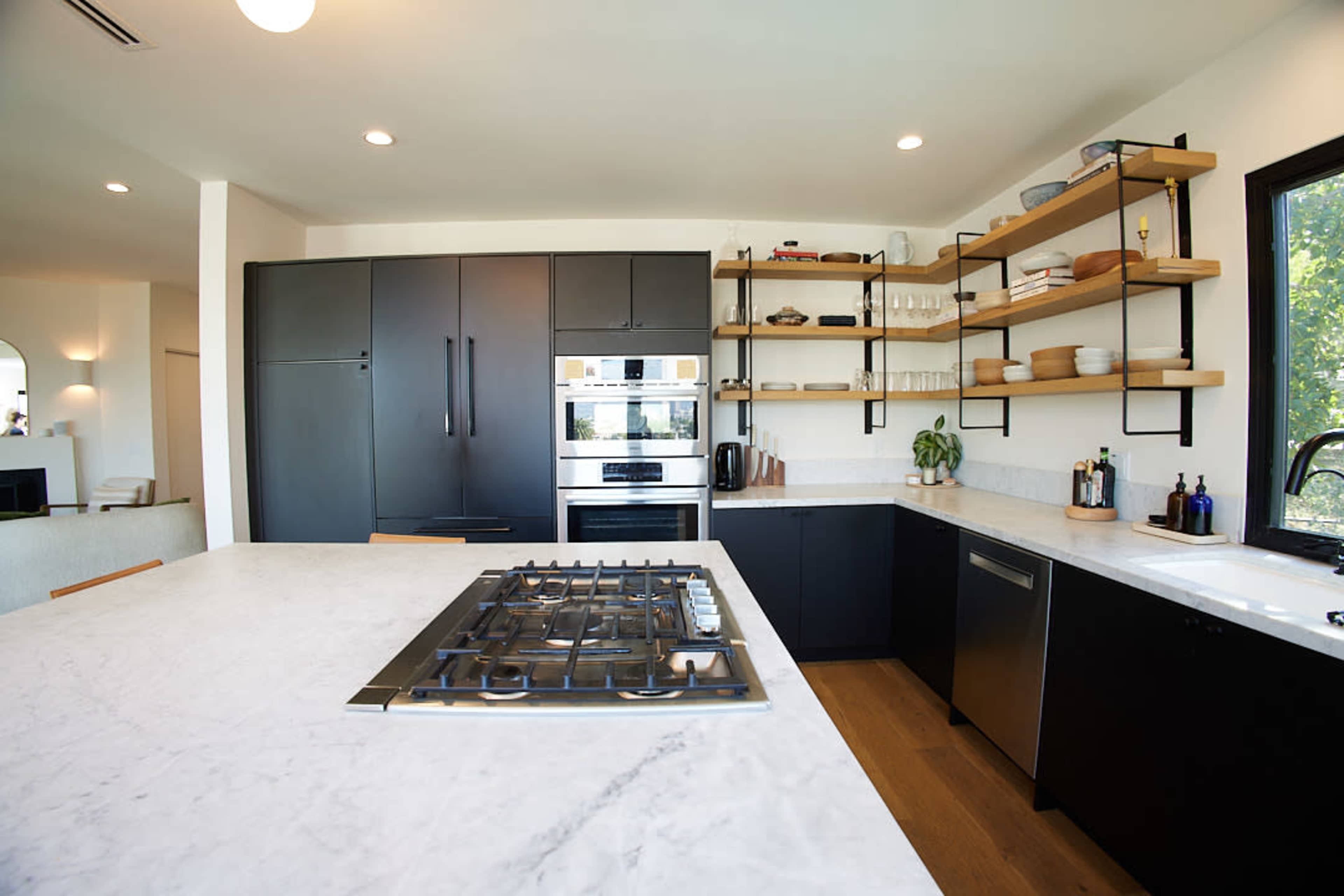 The image shows a modern kitchen featuring a marble countertop, black cabinetry, open shelving with dishware, and stainless steel appliances.