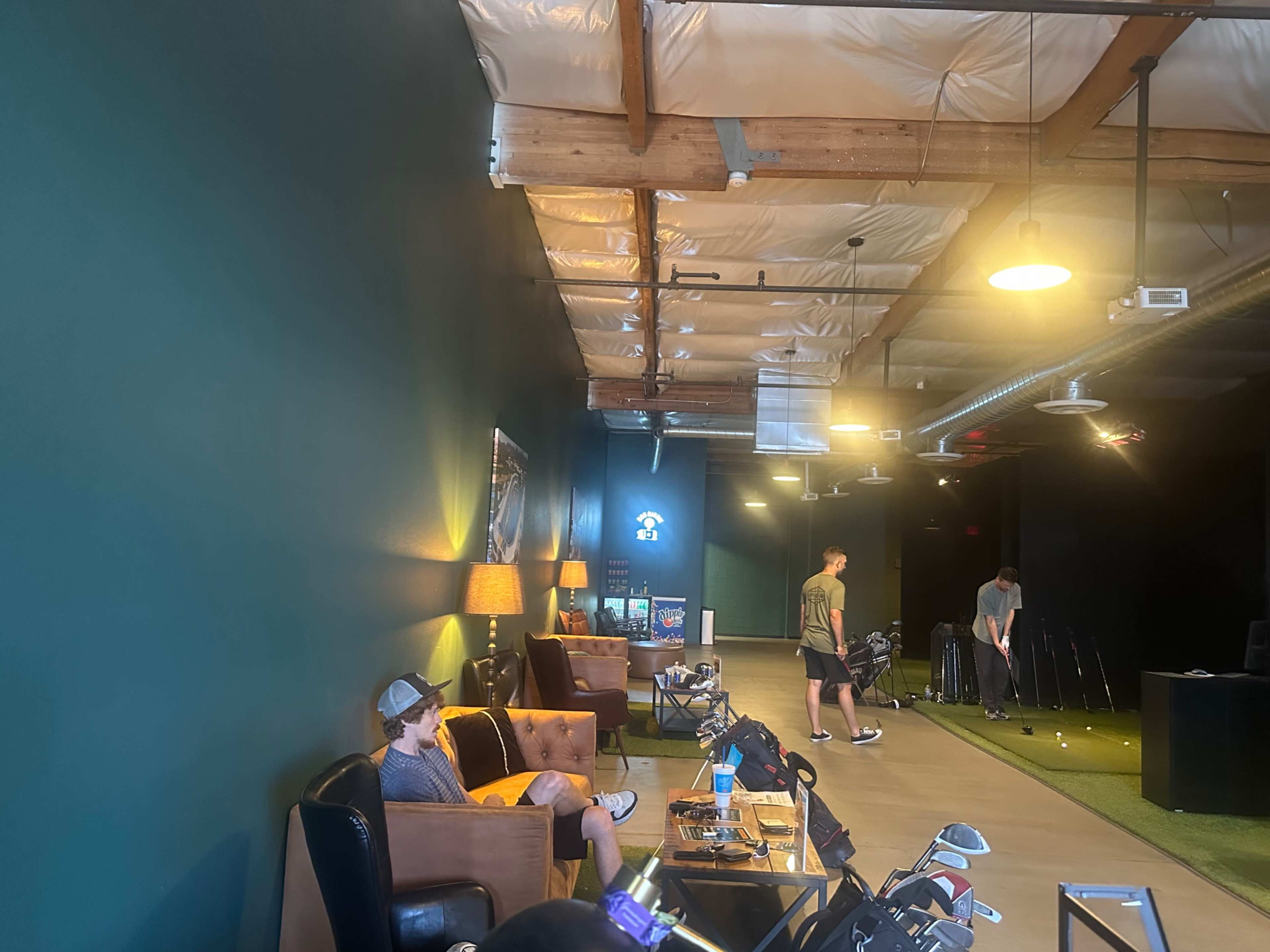 The image shows a spacious indoor golf practice facility with two men engaging in different activities, one sitting on a couch and the other practicing his swing.